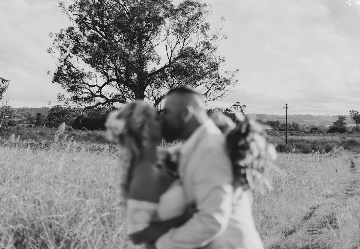 A black and white photo of a couple kissing in a rural outdoor setting with tall grass, trees, and a cloudy sky.