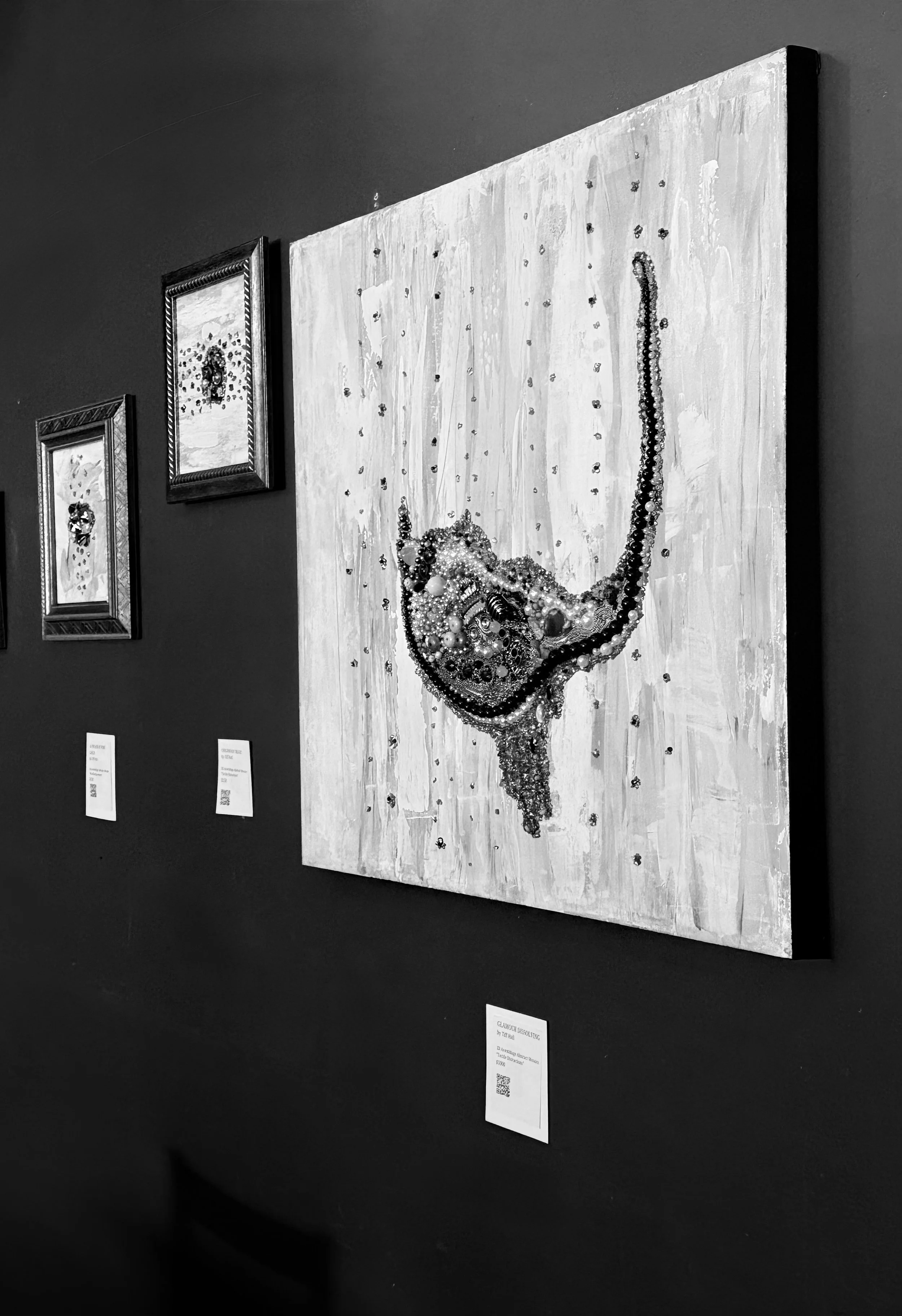 Black and white photo of artwork on a textured background, with two small informational plaques beneath and smaller framed pieces on the adjacent wall.