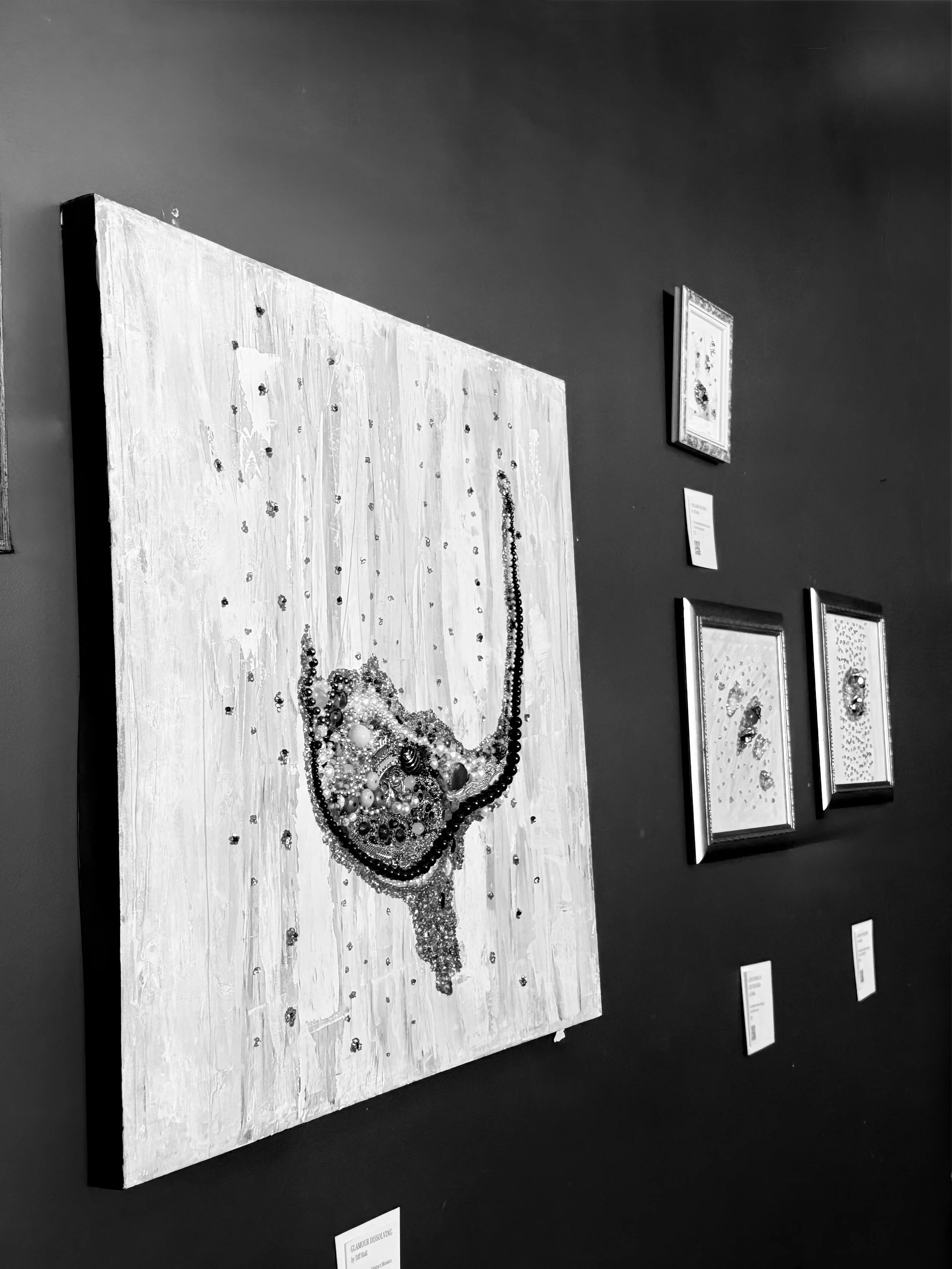 Black-and-white photograph of a mixed media art piece on display in a gallery, featuring various objects and beads arranged on a rectangular canvas.