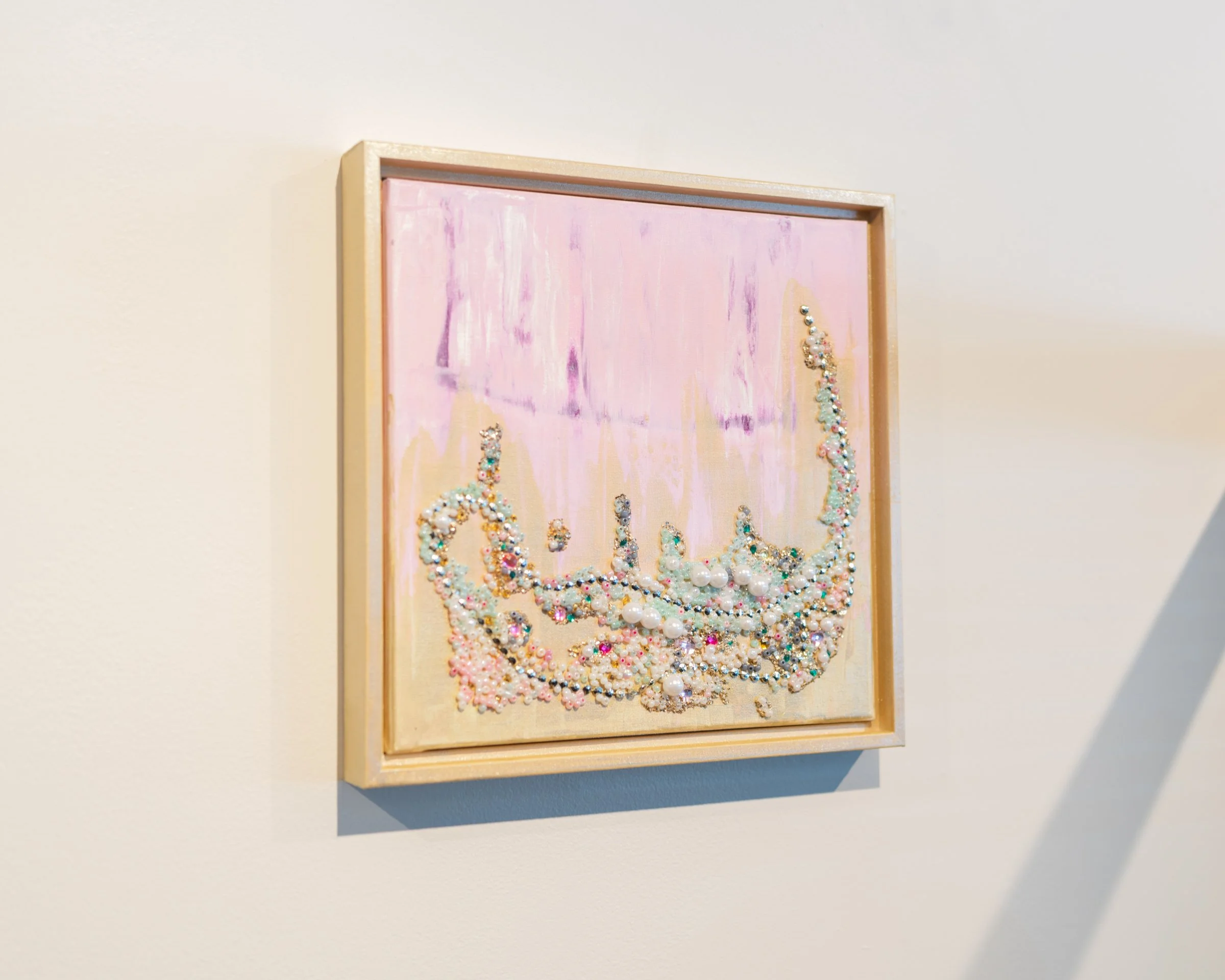 A framed artwork on a white wall featuring a pink background with a three-dimensional design of pearls and beads forming an elegant decorative pattern.