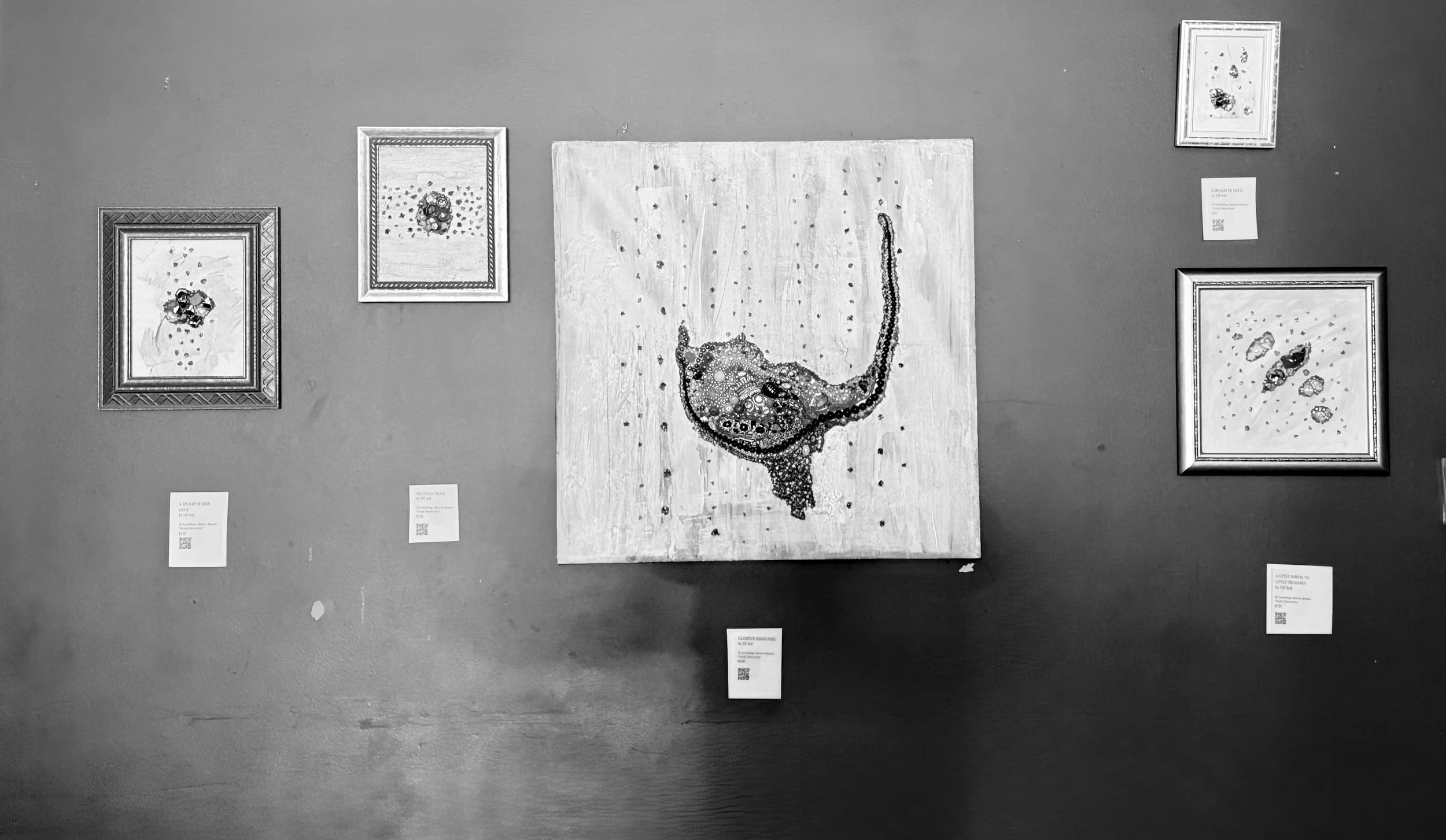 Black and white photograph of an art gallery wall displaying five framed artworks and one unframed piece. The artworks include abstract and figurative styles, with a central piece depicting a map or outline of a landmass formed by small dark objects.