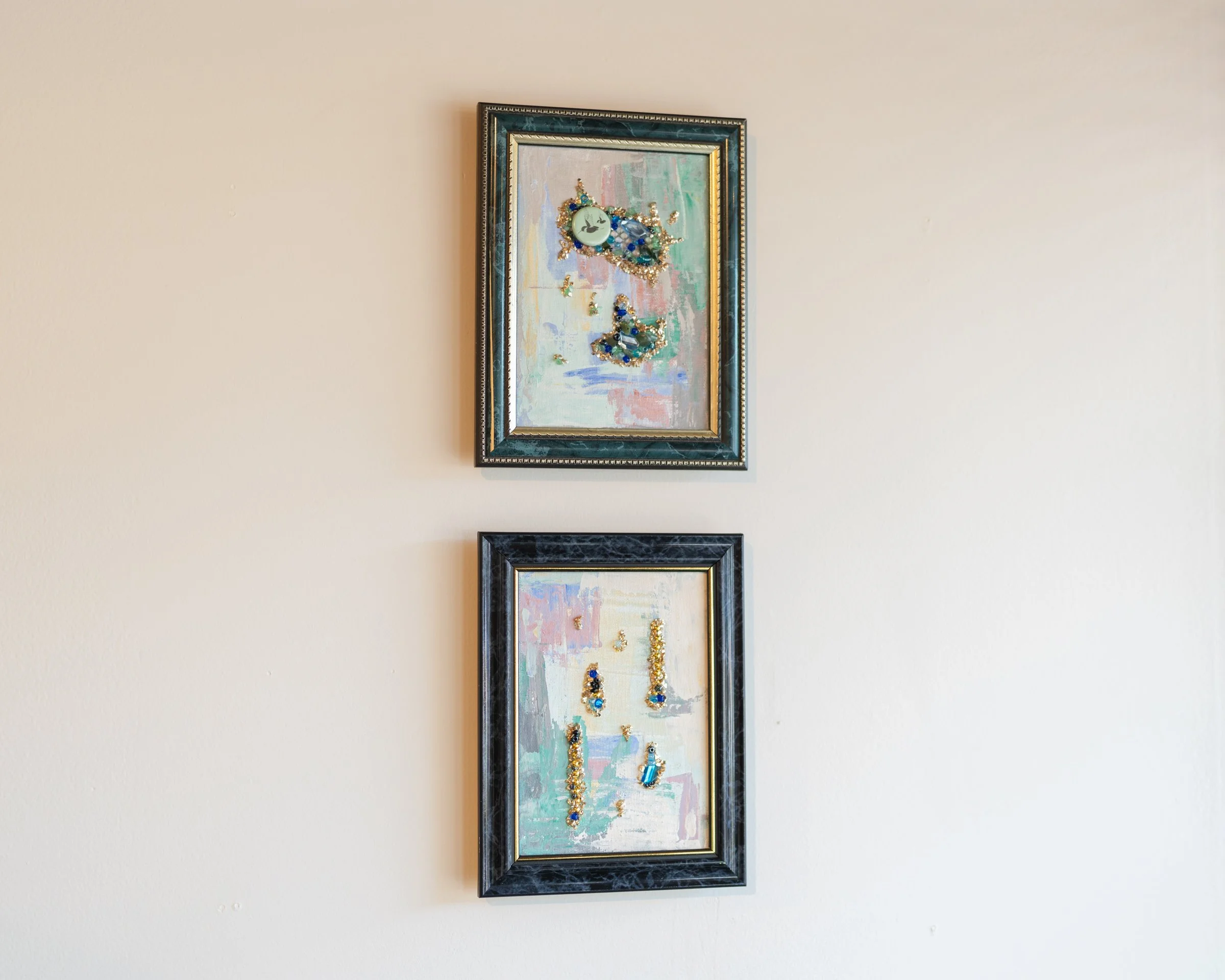 Two framed art pieces hanging on a beige wall, displaying jewelry such as earrings and necklaces, with an abstract pastel background.