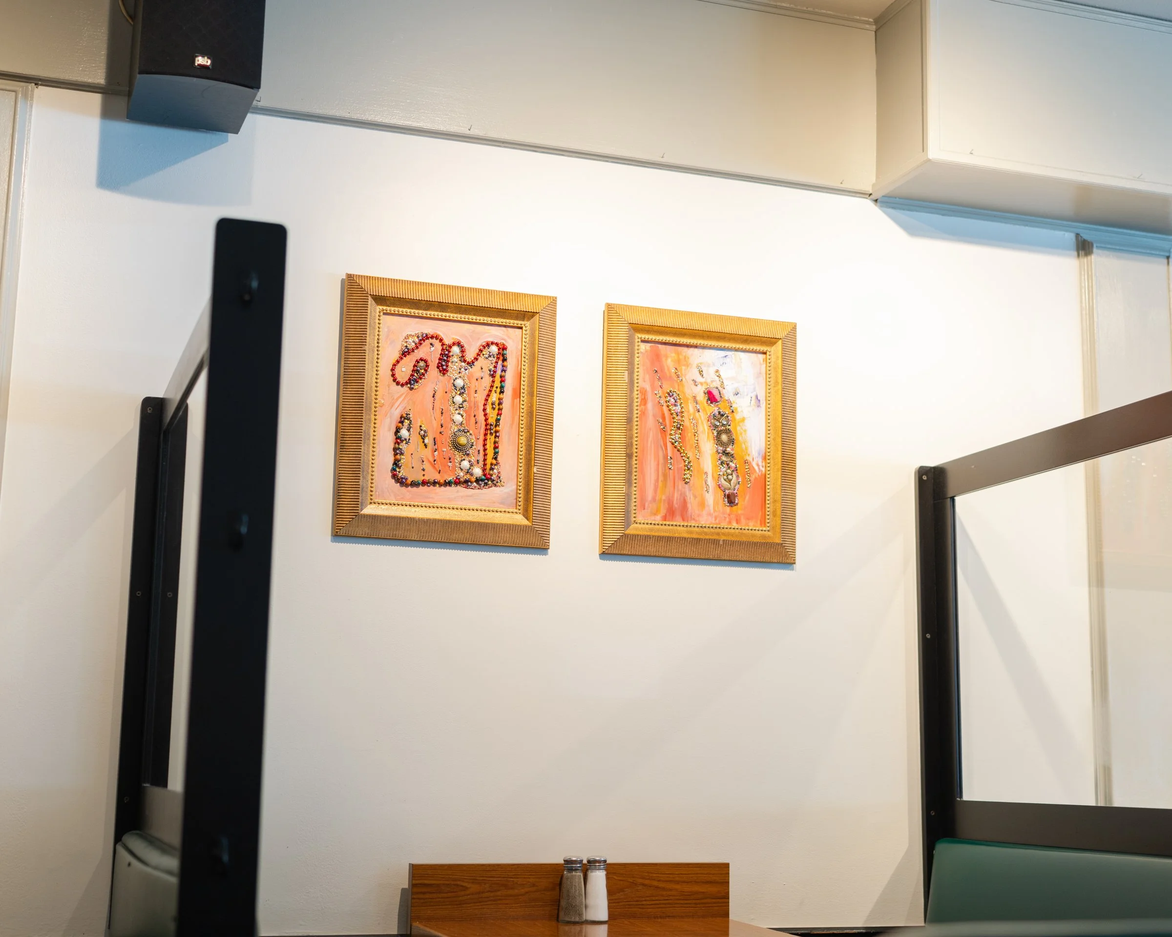 Two framed paintings with abstract designs hanging on a white wall in a room, with part of a black railing and a wooden table with salt and pepper shakers visible