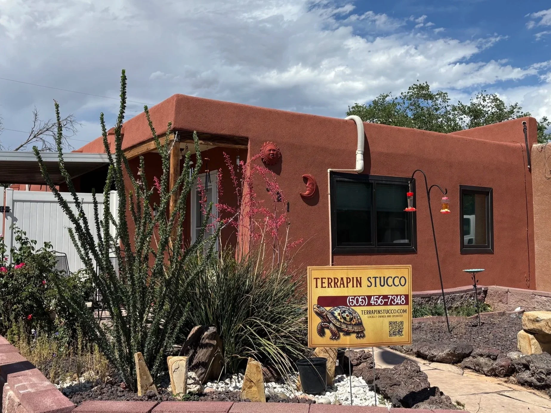 A building with terrapin-themed stucco exterior and tribal sun and moon wall decorations. There are desert plants in the foreground and a yellow sign with black text advertising terrapin stucco and the company's contact information.