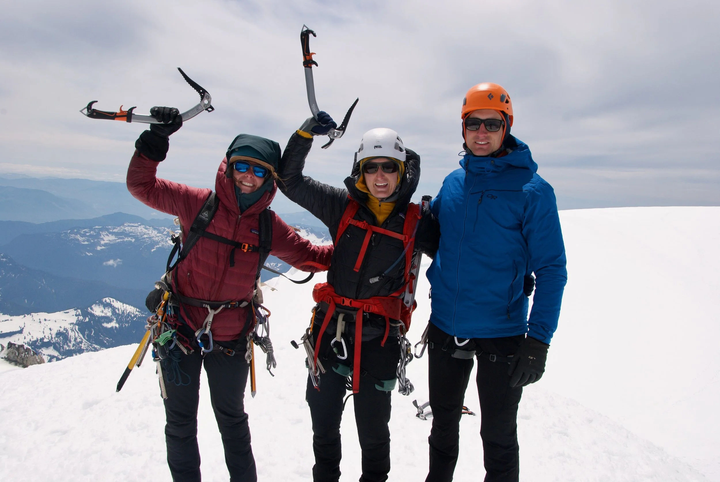 Three climbers on a snowy mountain peak, wearing winter gear, helmets, and sunglasses, holding ice axes, with mountain range in the background.