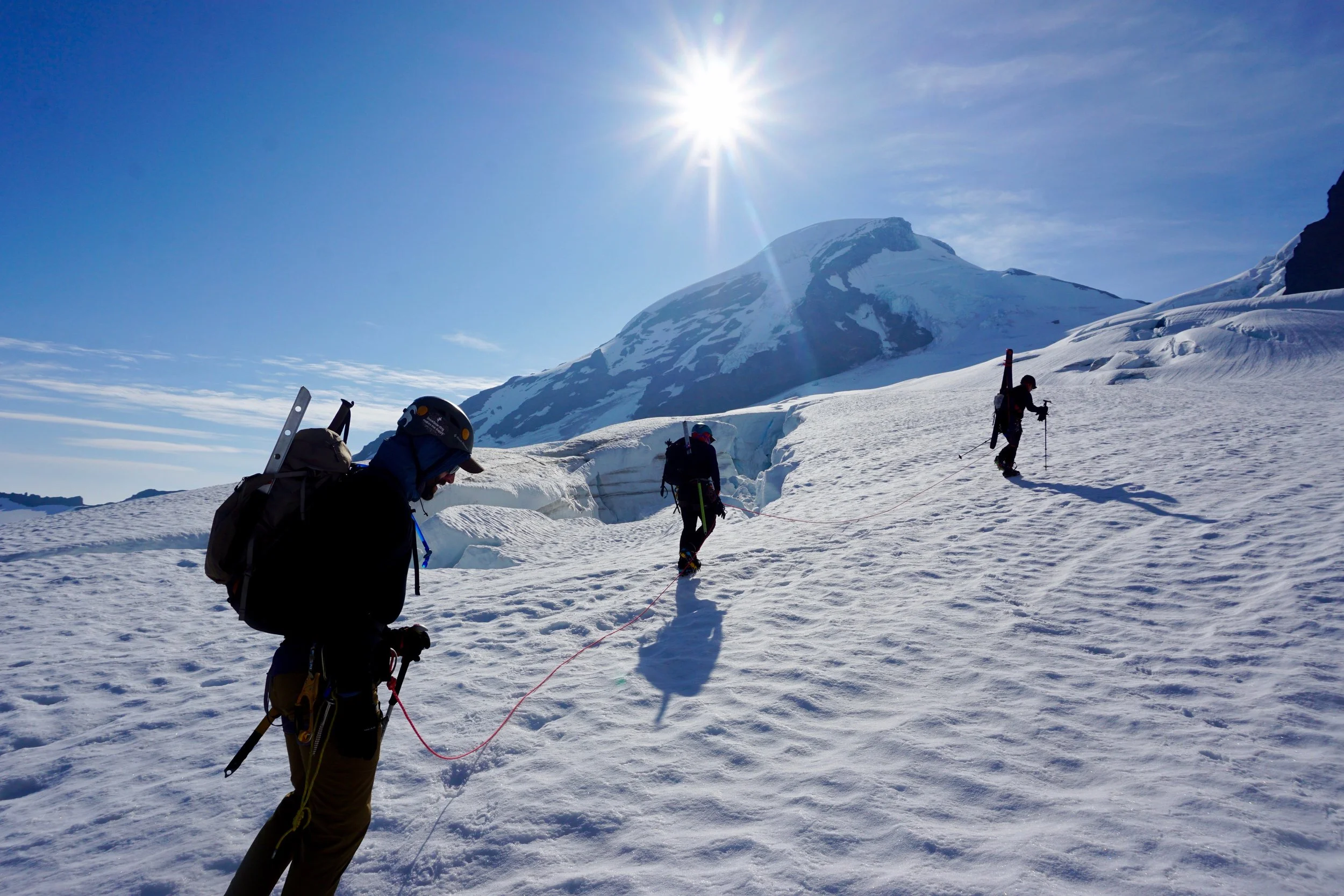 Three climbers ascending a snowy mountain slope in bright sunlight with a snow-covered mountain and clear blue sky in the background.