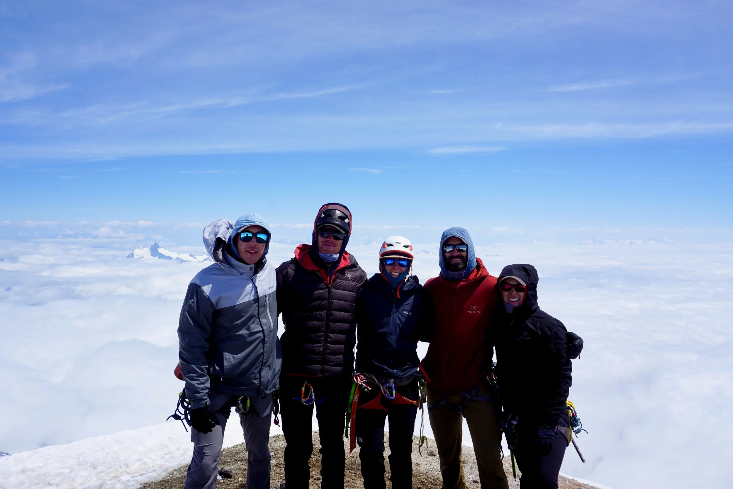 Group of five climbers on a snow-covered mountain summit with a vast cloudscape and a mountain peak in the background.