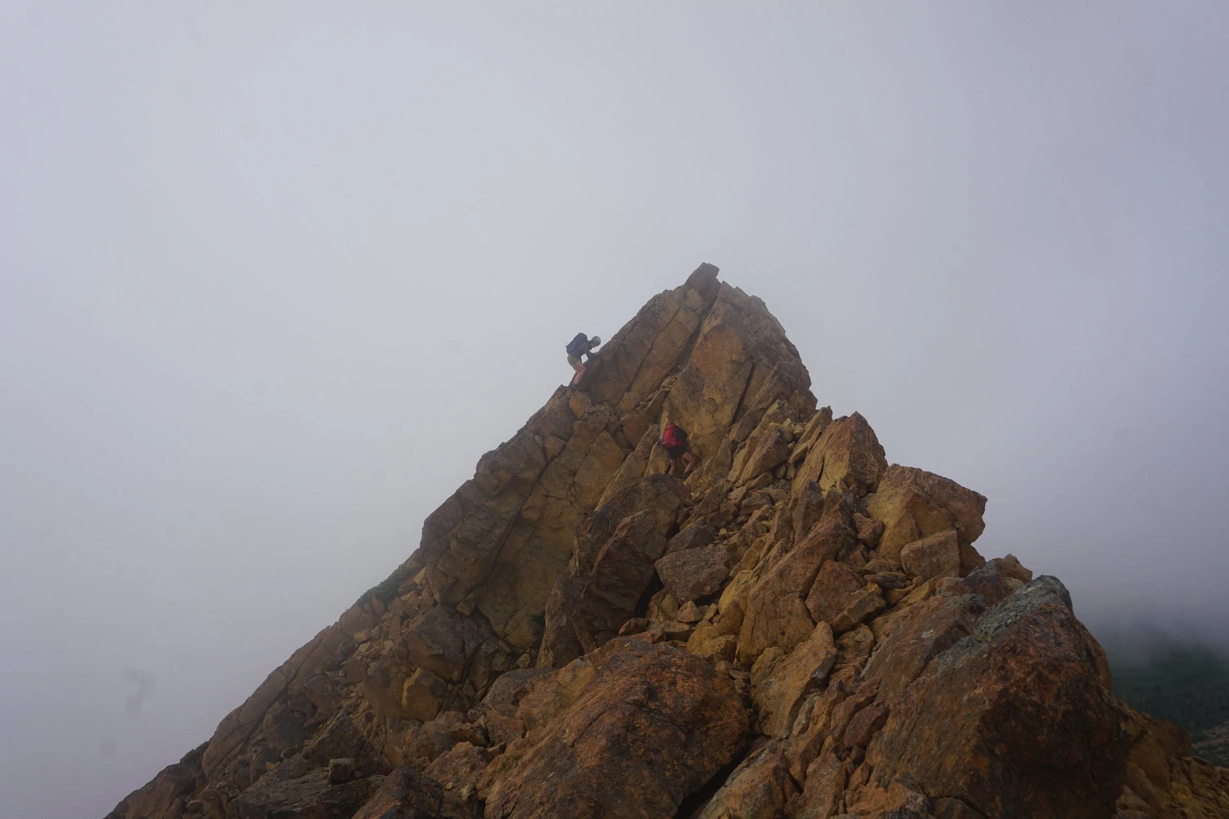 Two mountain climbers ascending a rocky, steeper section of a mountain with foggy, overcast sky.