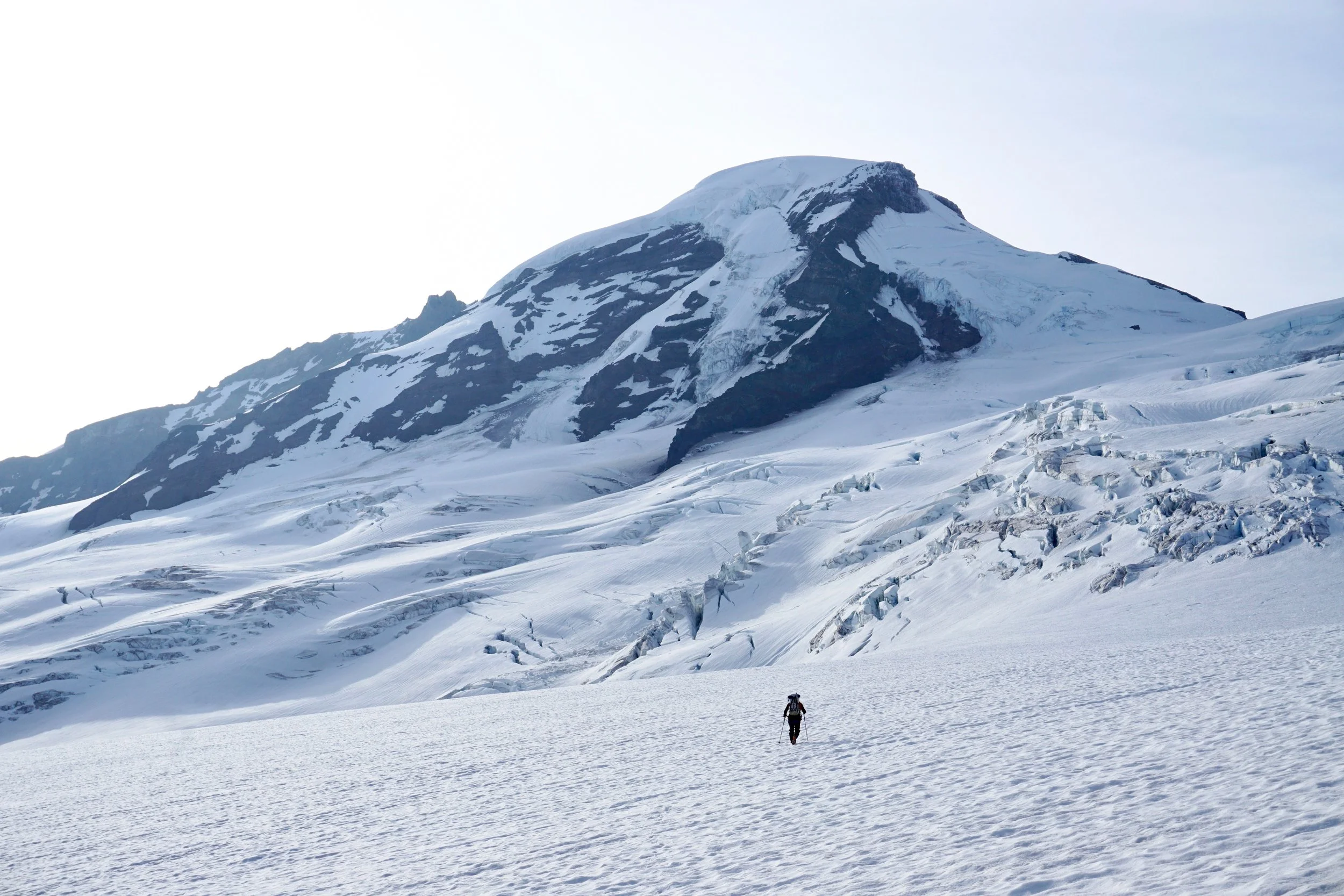 A person hiking on a snow-covered glacier with a large snow-capped mountain in the background.