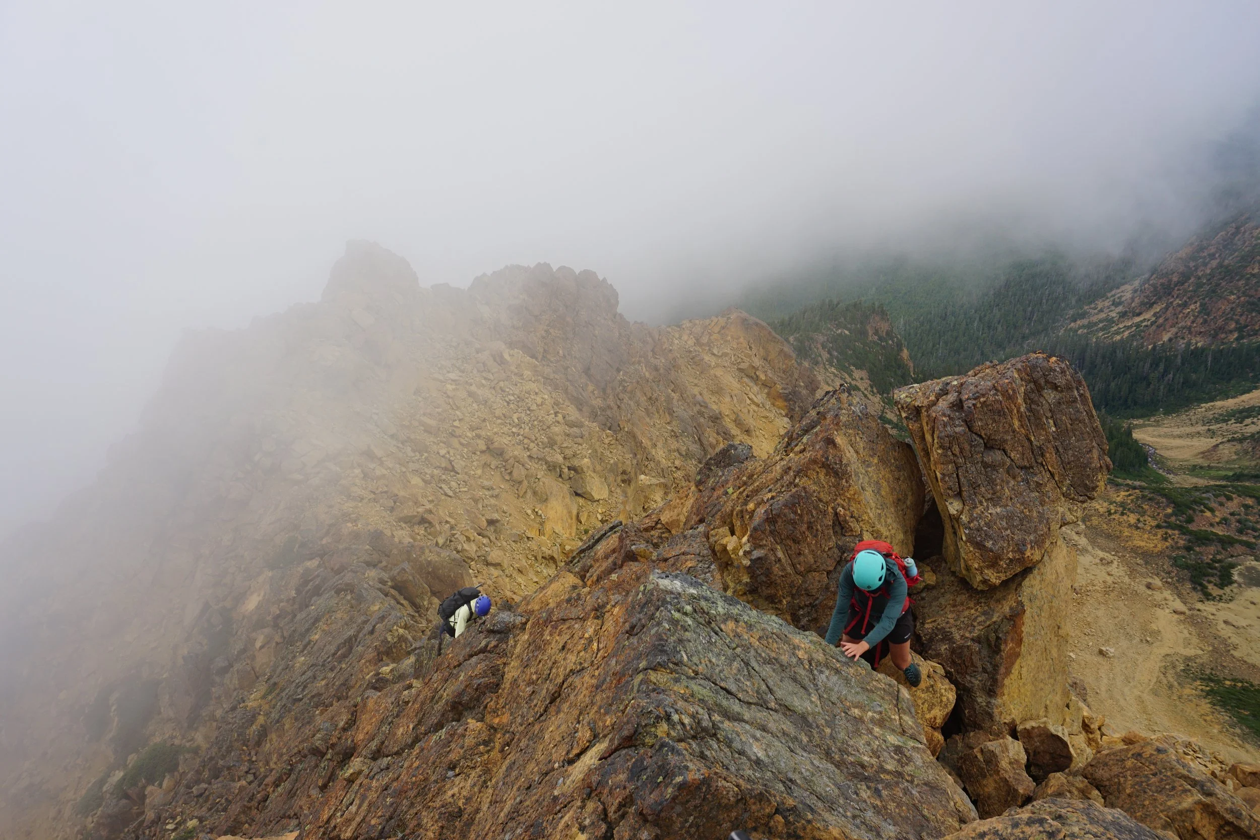 Two climbers ascending a steep rocky mountain with fog and clouds in the background.