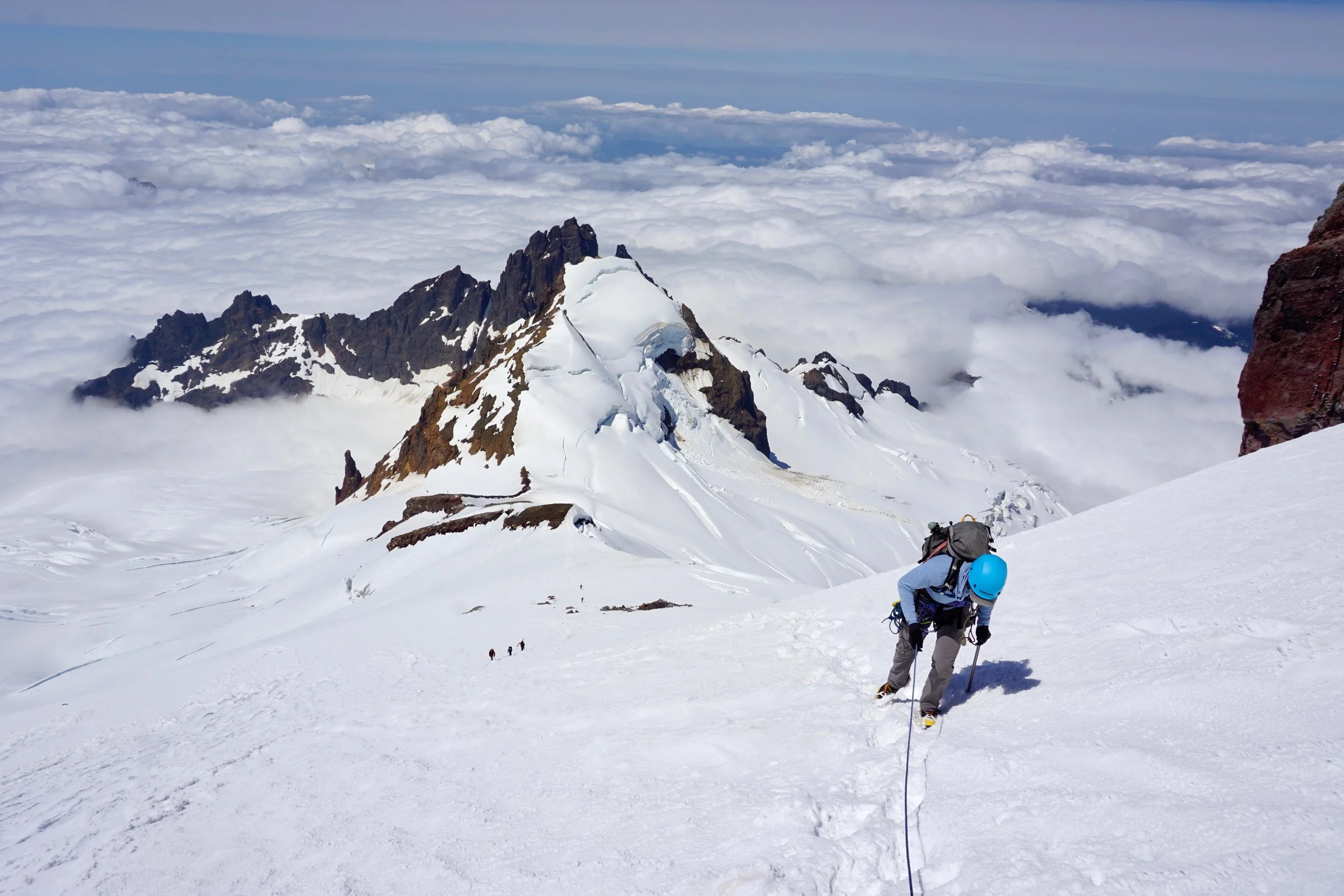 A mountaineer climbing a snowy, steep mountain slope with a rope, wearing a blue helmet and backpack, with more climbers visible in the distance and a mountain peak above a sea of clouds.