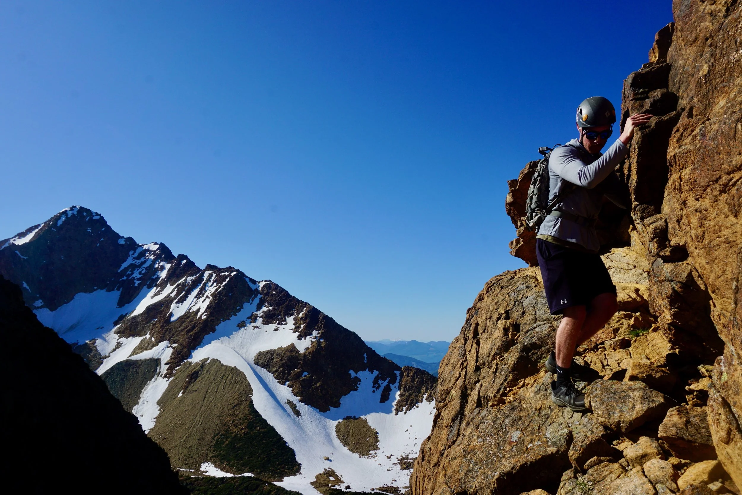 A man wearing a helmet, sunglasses, and outdoor gear climbs a rocky mountain trail with snow-capped peaks in the background.