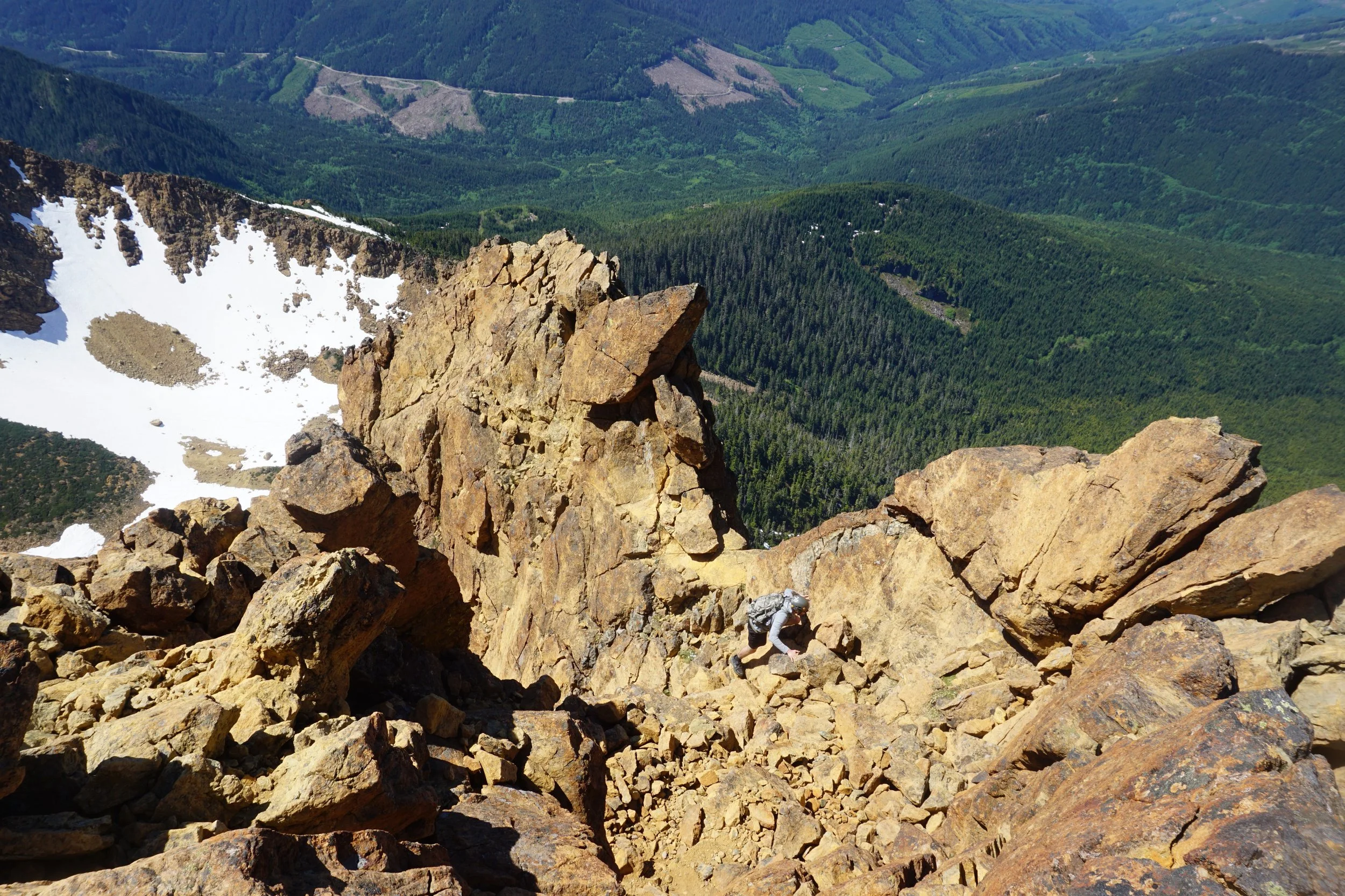 A person climbing over large rocks on a mountain trail with a scenic valley and snow patches below.