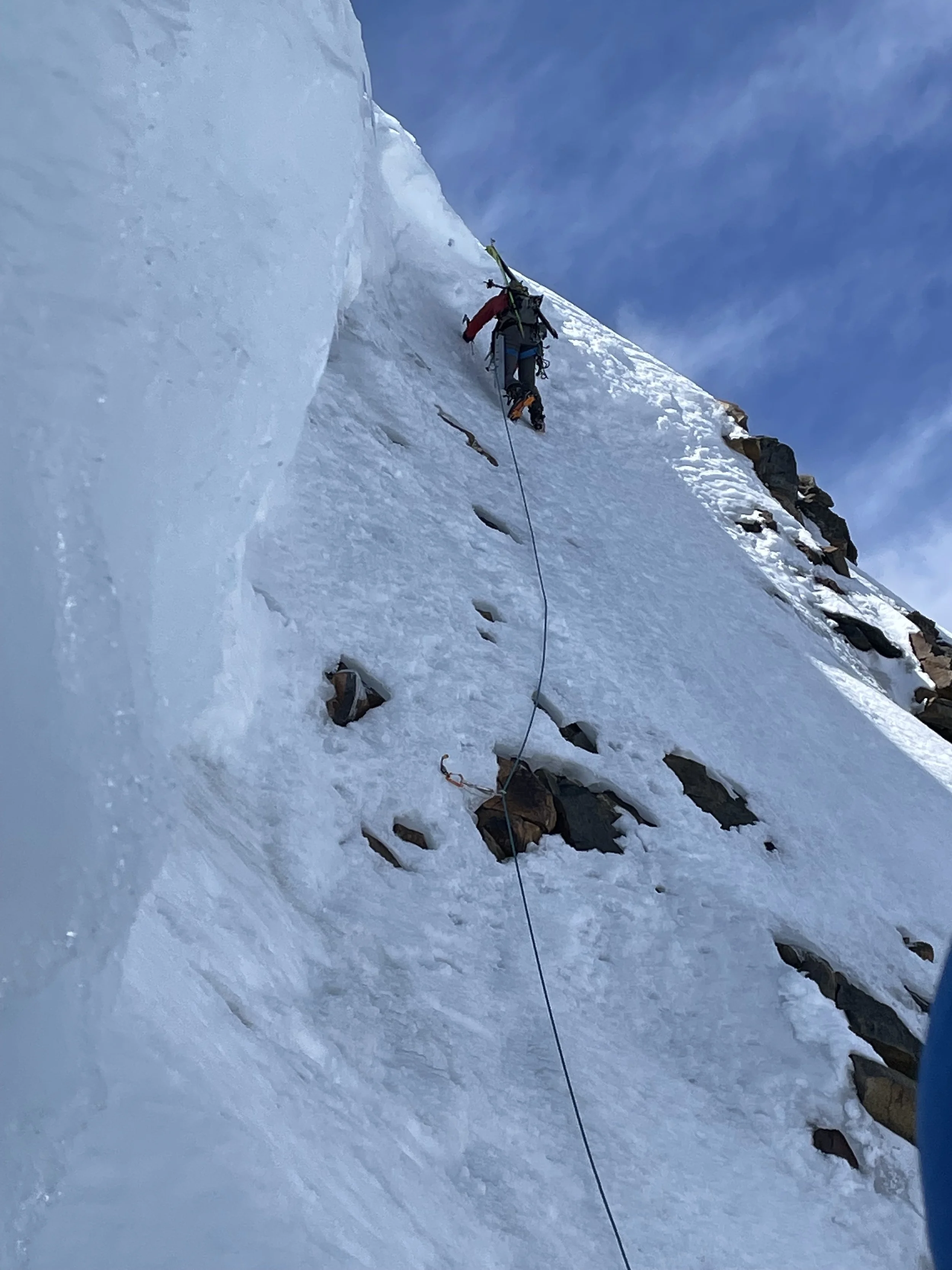 A climber ascending an icy mountain face with climbing gear, wearing a red and blue jacket, and a helmet, using an ice axe and crampons on the snow and ice surface.