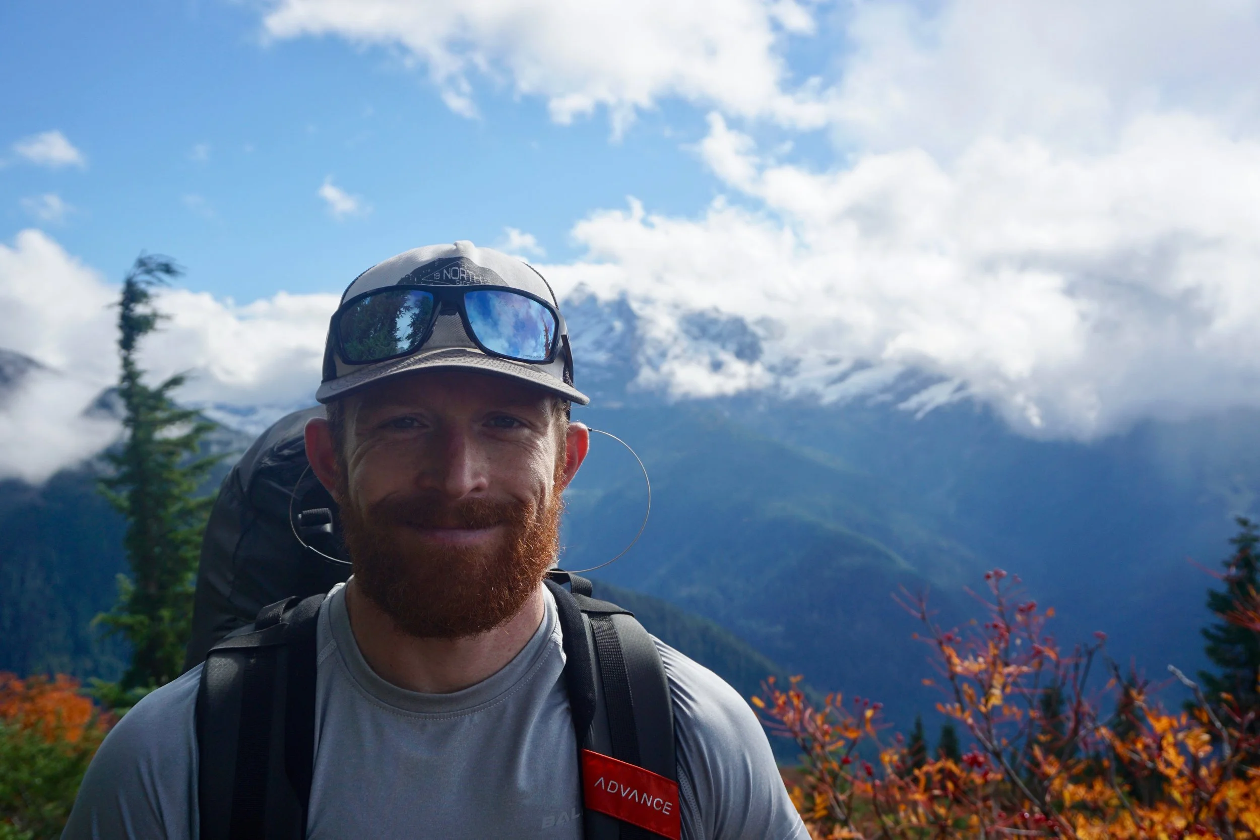 A man with a red beard, wearing a gray cap with sunglasses, a gray T-shirt, and a backpack, stands in front of a mountainous landscape with snow-capped peaks, clouds, and greenery.