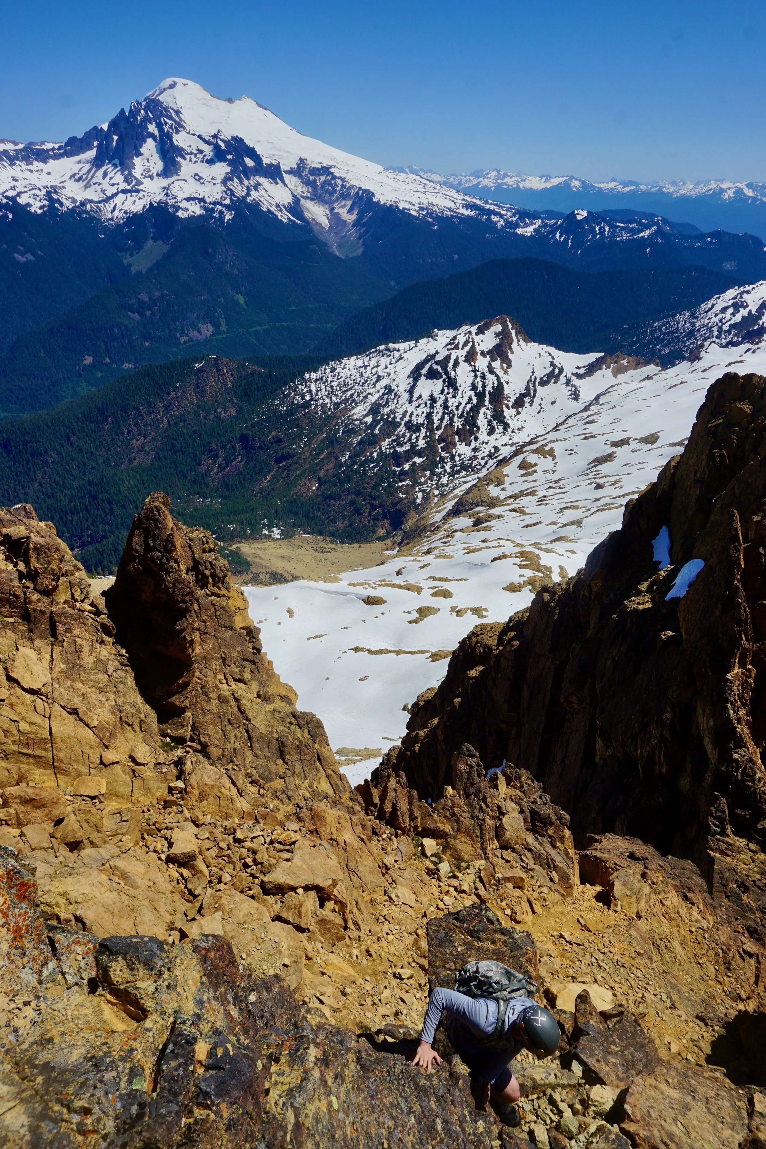A mountain climber wearing a helmet and backpack ascending a rocky slope on a rugged mountain trail, with snow-capped peaks and green valleys in the background.