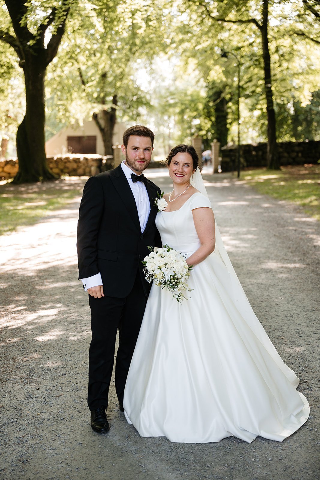 Brudepar i svart dress og hvit brudekjole med blomsterbukett på en skogkledd grusvei