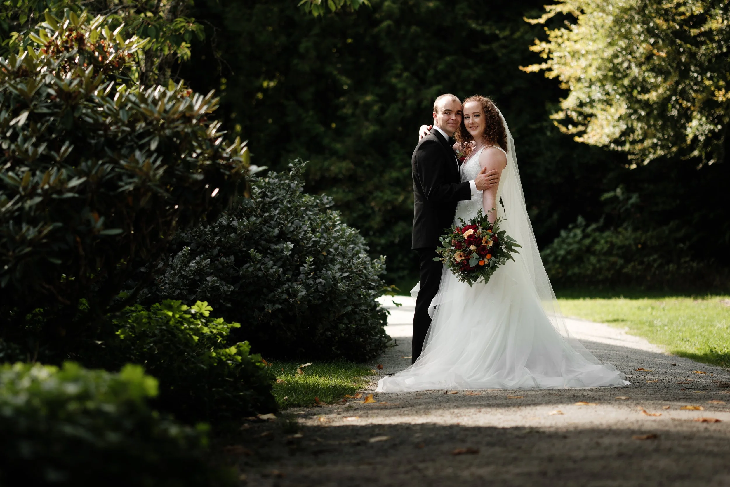Et brudepar står sammen på en sti i en grønnhage, kledd i brudekjole og smoking, med blomsterbukett.