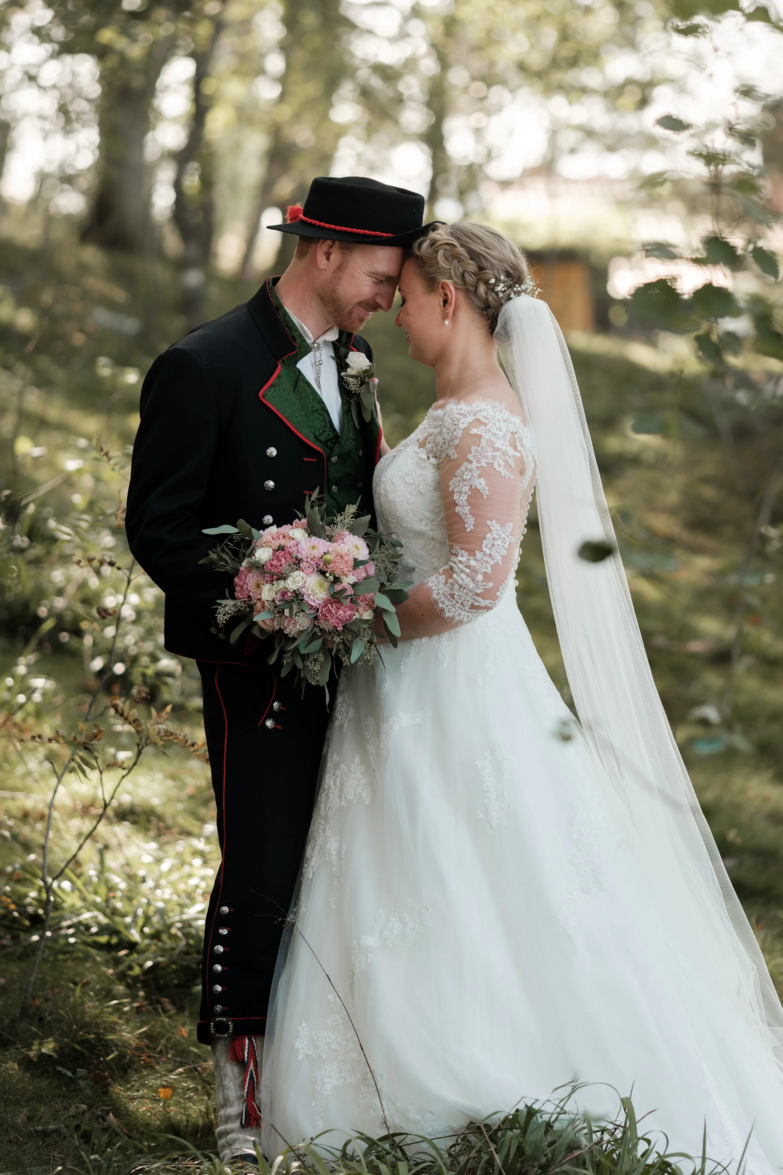 Et brudepar står nære sammen i en skog, kledd i tradisjonelle norske folkedrakter, med hodet tilbrakt og ansikt vendt mot hverandre, mens den brud glemmer å bære brudebuketten.