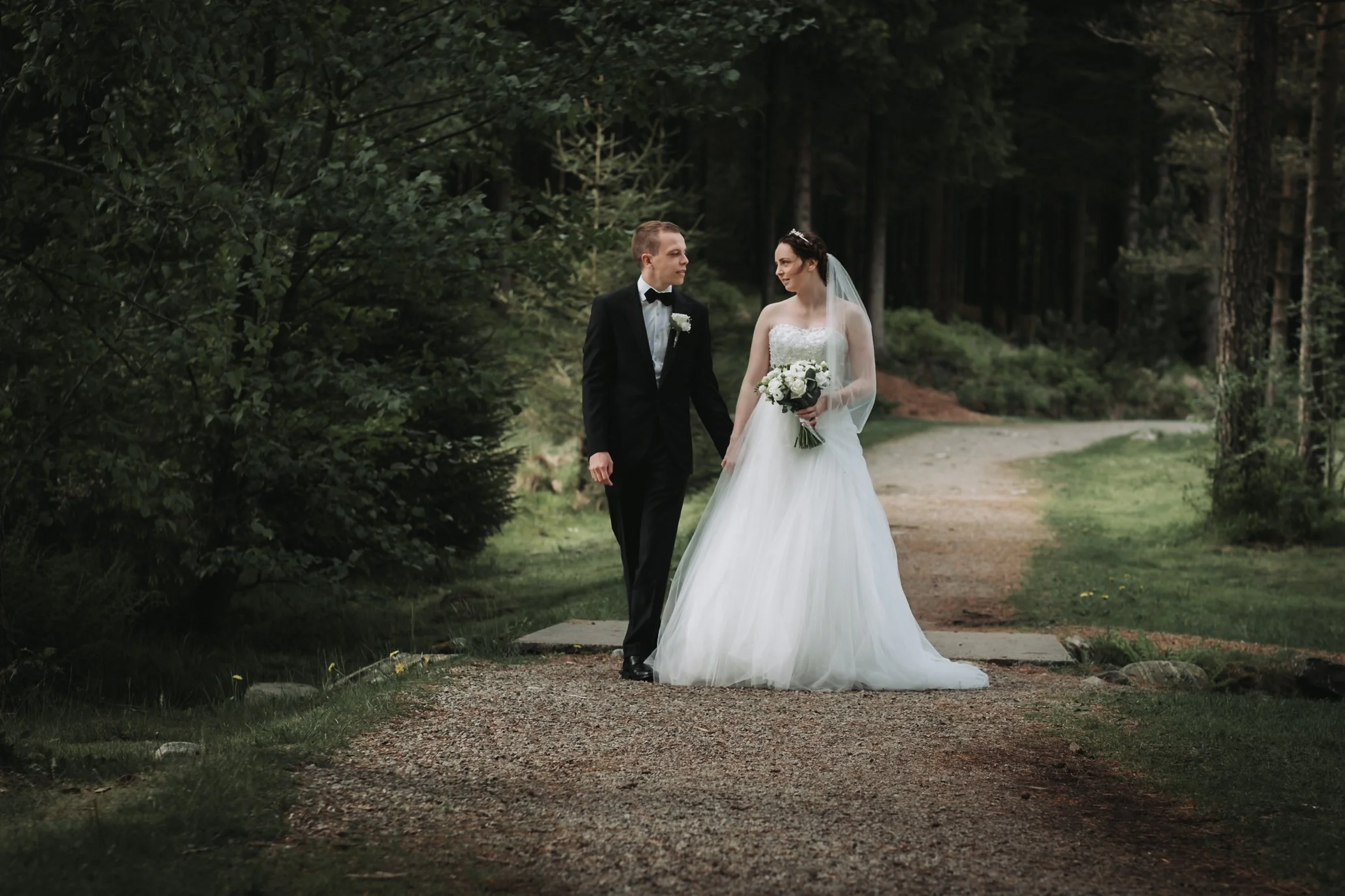 Et brudepar går hånd i hånd langs en skogssti, med bruden i en hvit brudekjole og brudgommen i en svart dress, omgitt av trær og grønt landskap.
