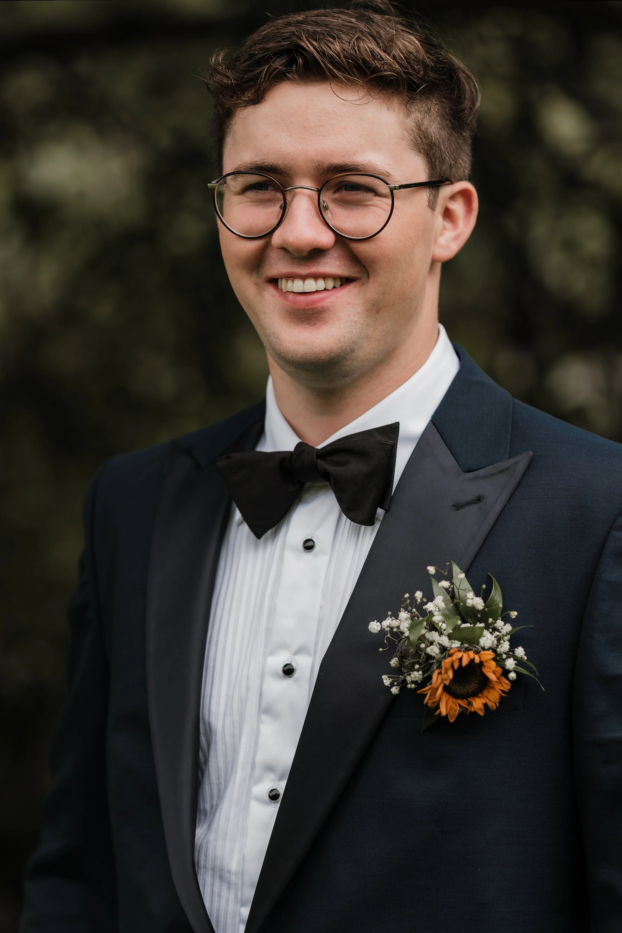 Ung mann i smoking med svart slipser og hvit skjorte, har brunt krøllete hår, bærer briller, smiler, har en bukett med solsikke og små hvite blomster i jakkelommen.