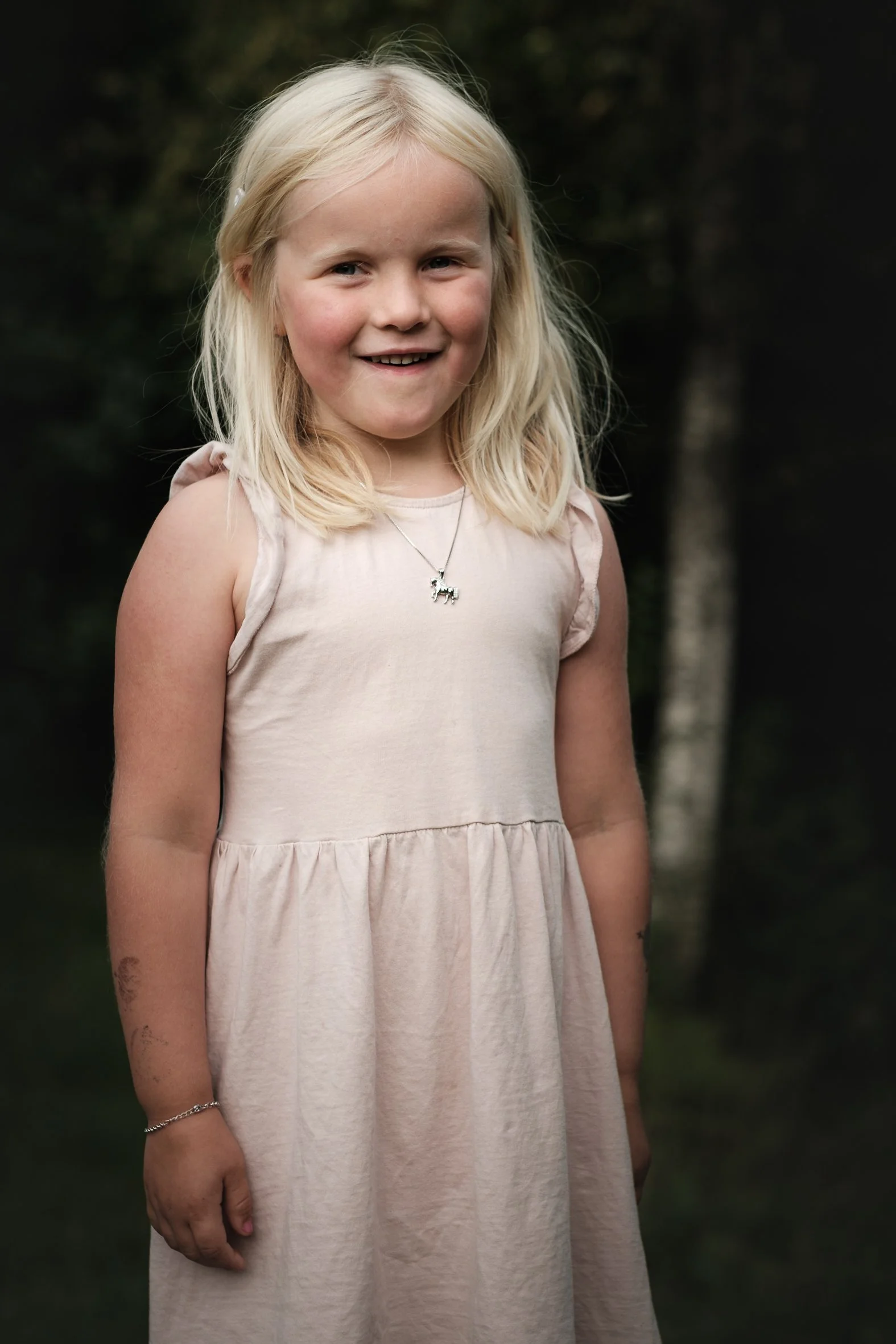 Smilende blond jente i lys kjole med halskjede, fotografert utendørs foran en mørk skog