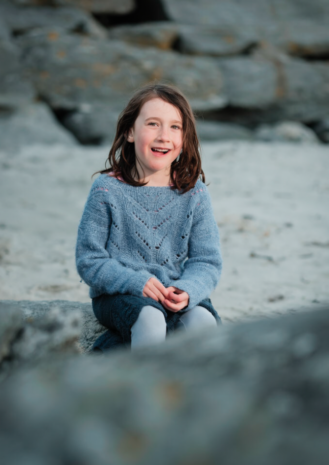 Liten jente sitter på en stein ved stranden, smiler, med steiner og en klippevegg i bakgrunnen.