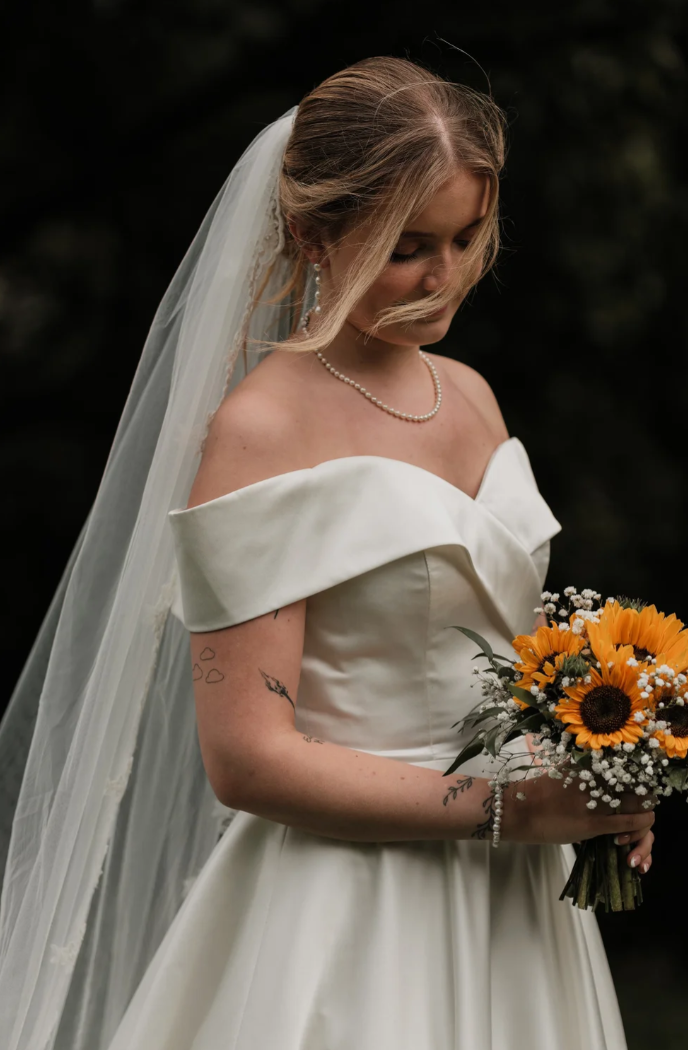 Brud i hvit off-shoulder-brudekjole med slør, perlekjede og bukett med oransje blomster