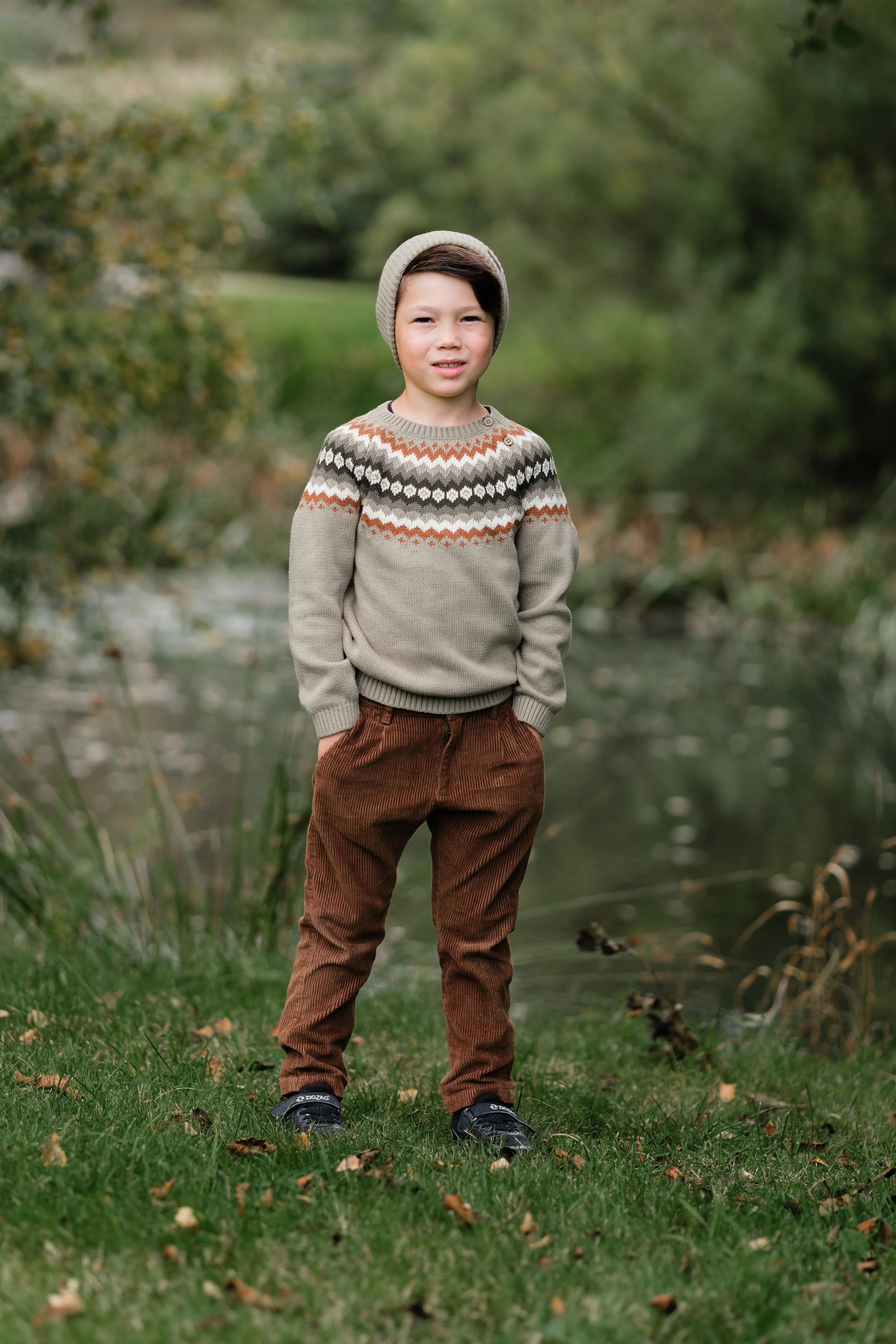 En gutt står på gressplen ved en elv, iført en strikkejakke, brune cordbukser og en beige lue, med natur i bakgrunnen.