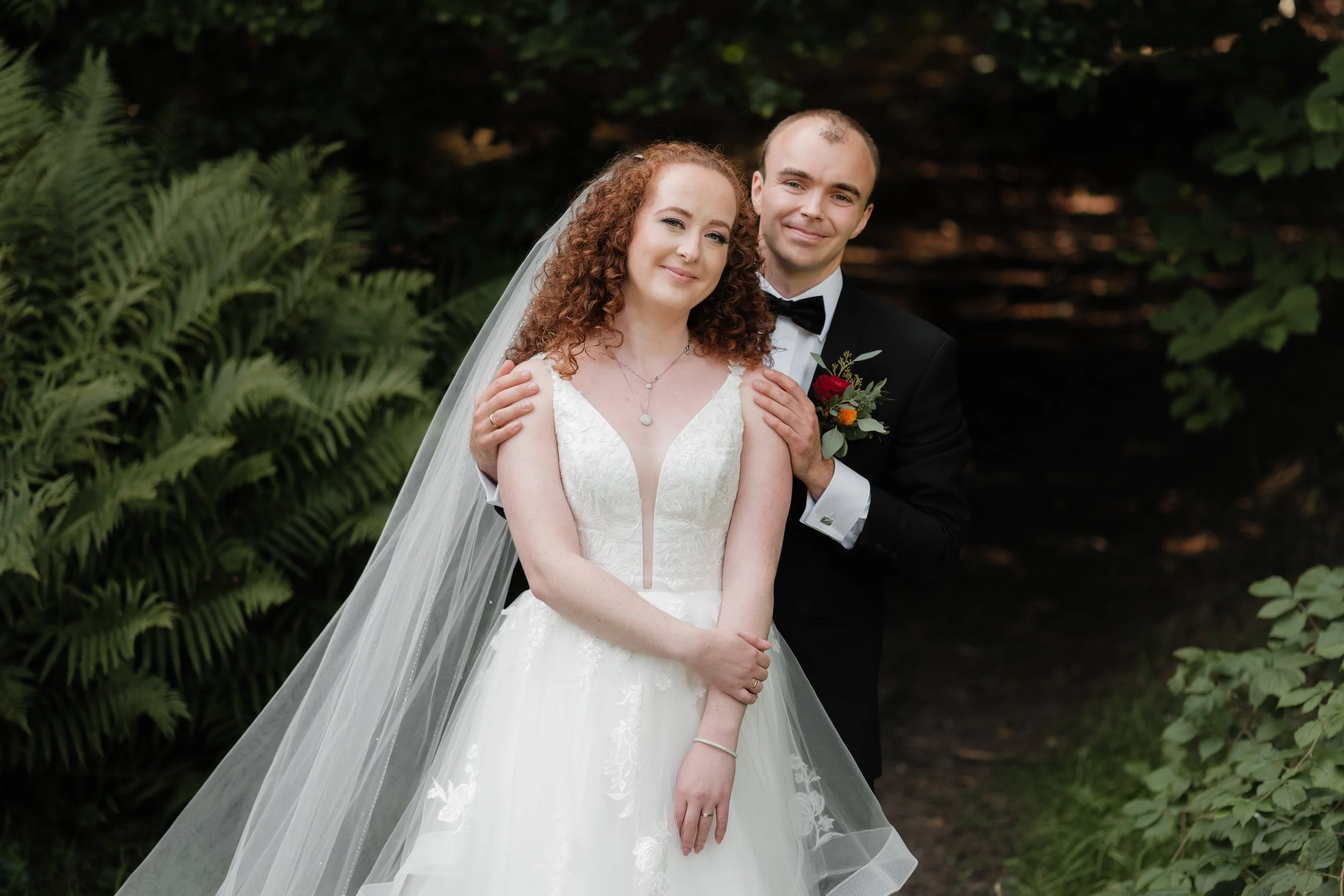 Et brudepar som står sammen utendørs, med grønne planter og trær i bakgrunnen.