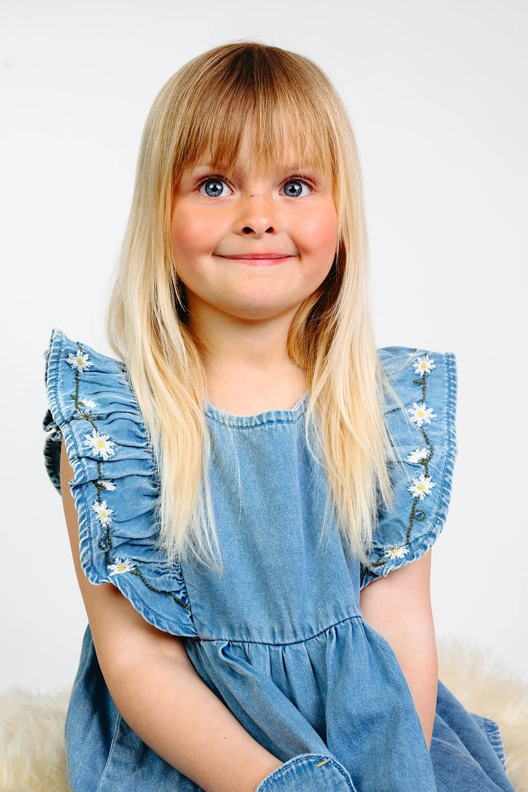 Smilende blond jente i blå denimkjole med blomstrete volanger, fotografert mot lys bakgrunn.