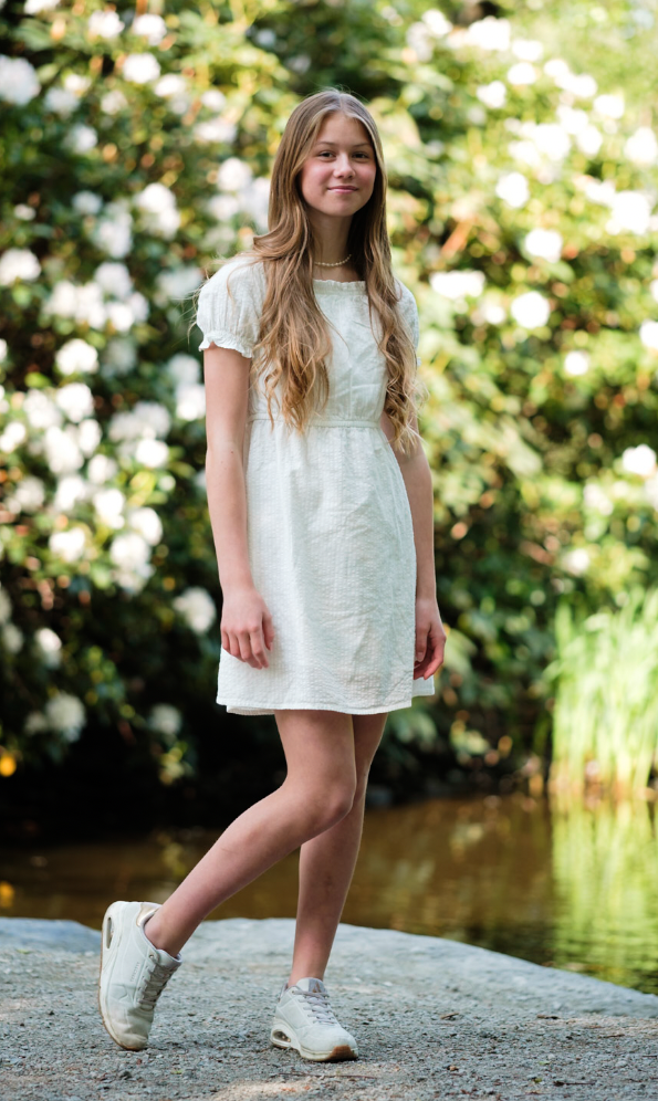 En ung jente står på en stein ved en liten innsjø, omgitt av grønne busker og hvite blomster.