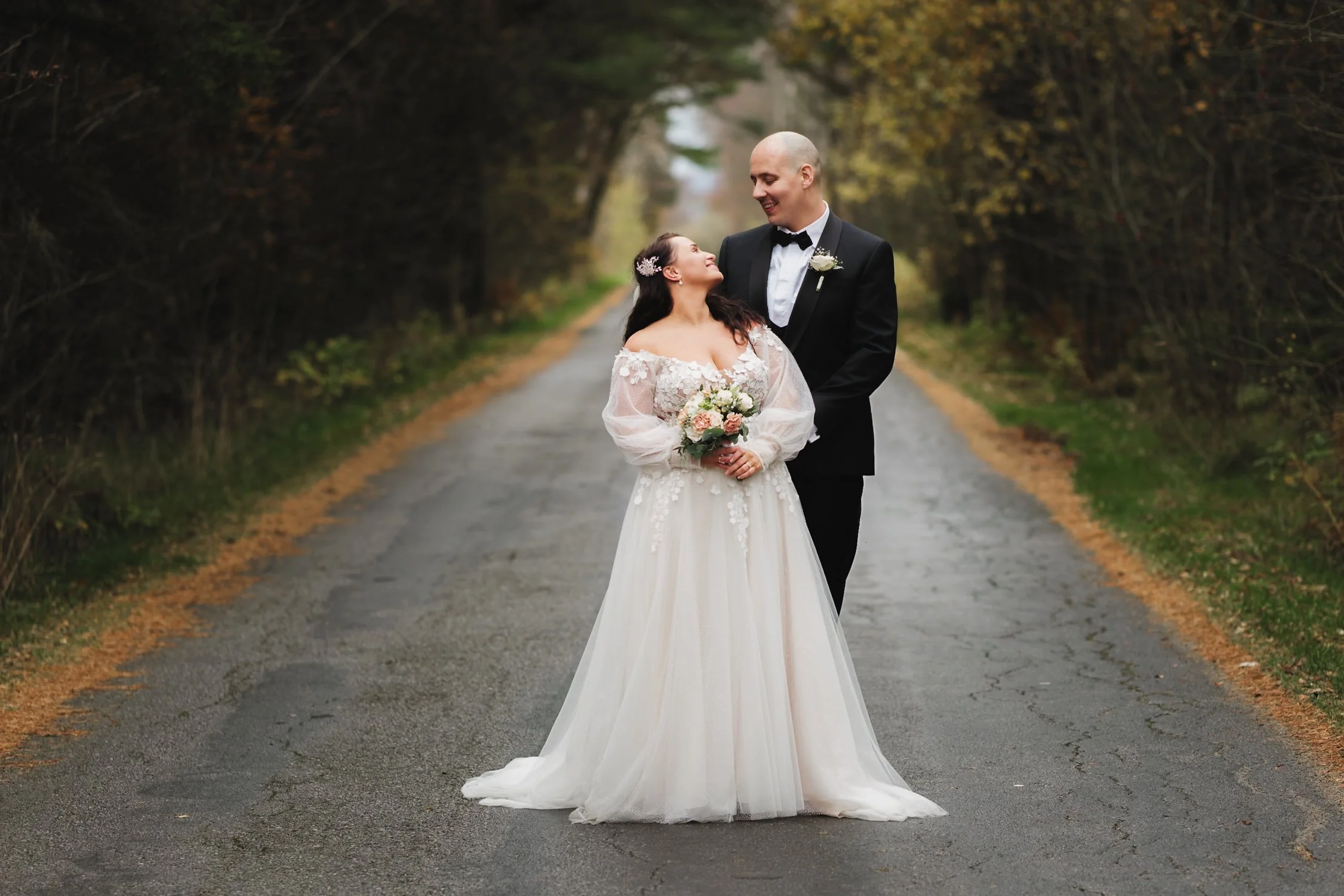 Bryllupspar som står på en villvei omgitt av trær med høstfarger, kledd i brudekjole og dress og ser på hverandre med kjærlighet.
