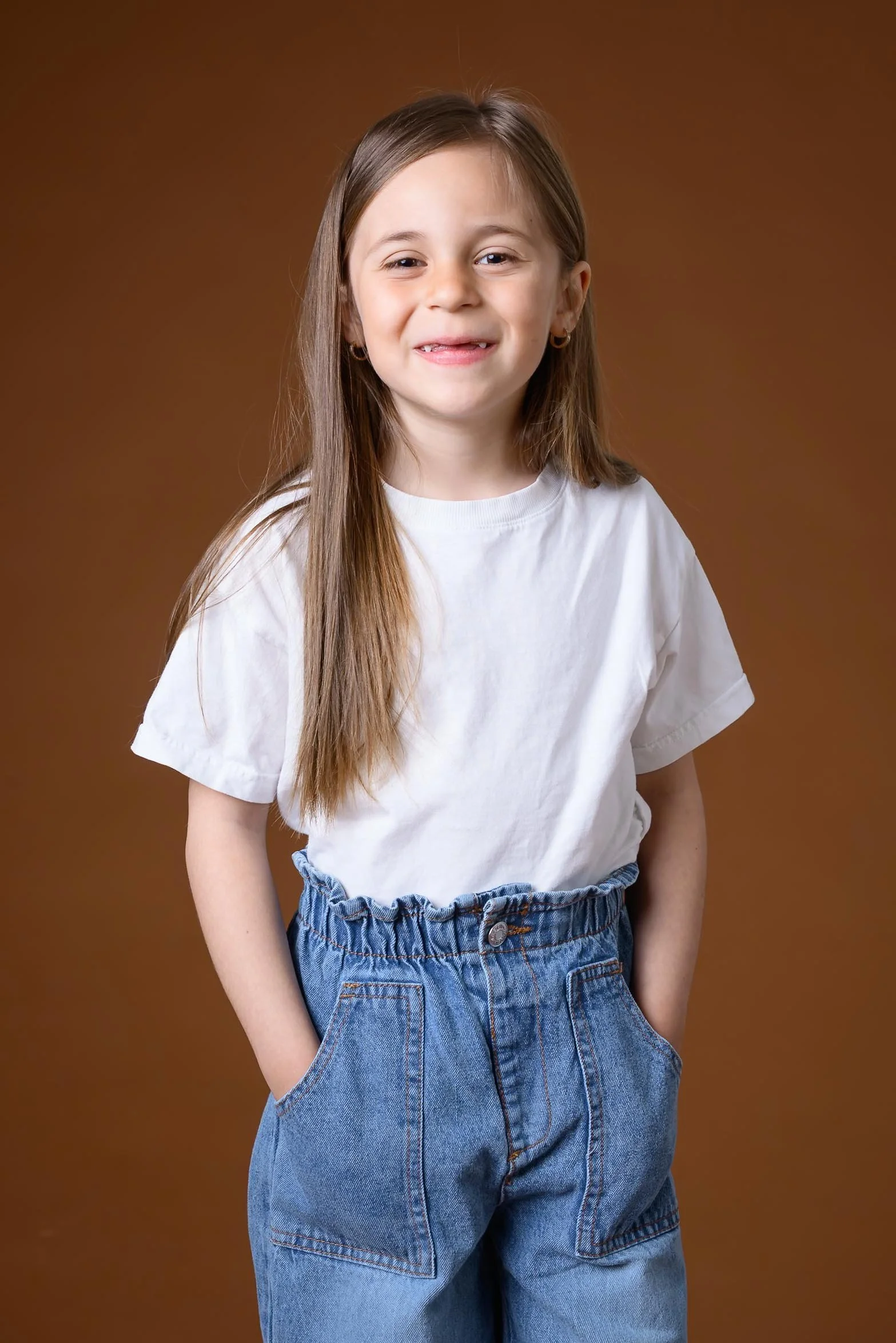 Ung jente med langt, brunt hår som smiler, iført hvitt T-skjorte og jeans, stående mot en brun bakgrunn.