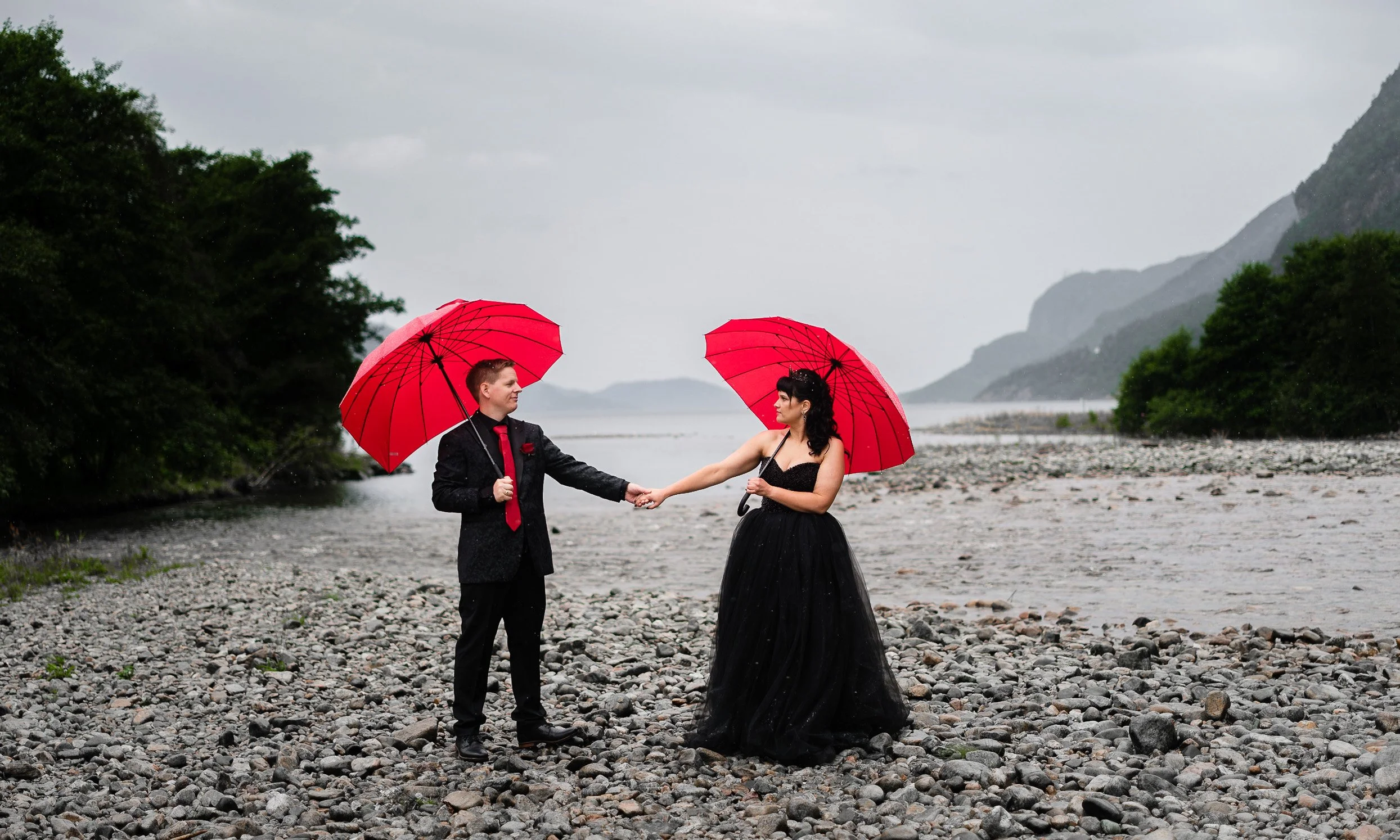Et kledd par i formelle klær holder hverandre i hendene under paraplyer ved en grusstrand, med en elv og fjell i bakgrunnen i regnvær.