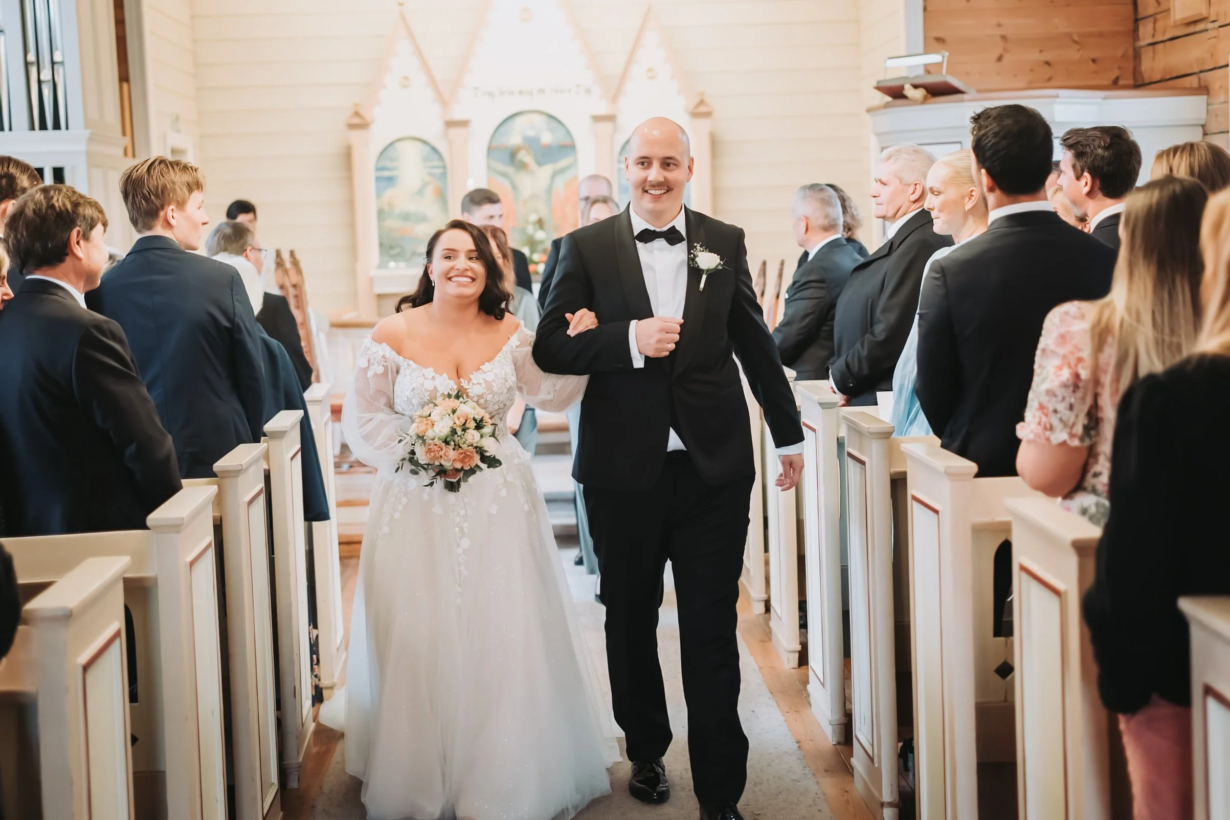 Et brudepar går ned midtgangen i en kirke, omgitt av gjester. Bruden holder en bukett blomster, og brudgommen har på seg en smoking.