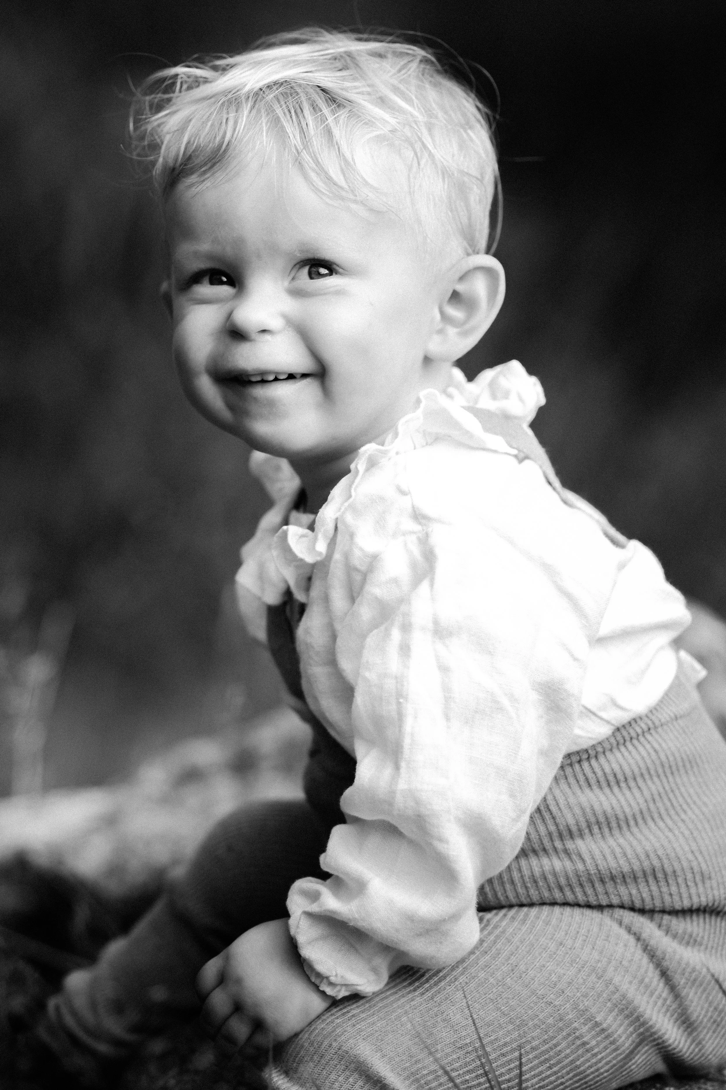 Smilende baby med lyst hår i hvit kjole, fotografert i svart-hvitt.