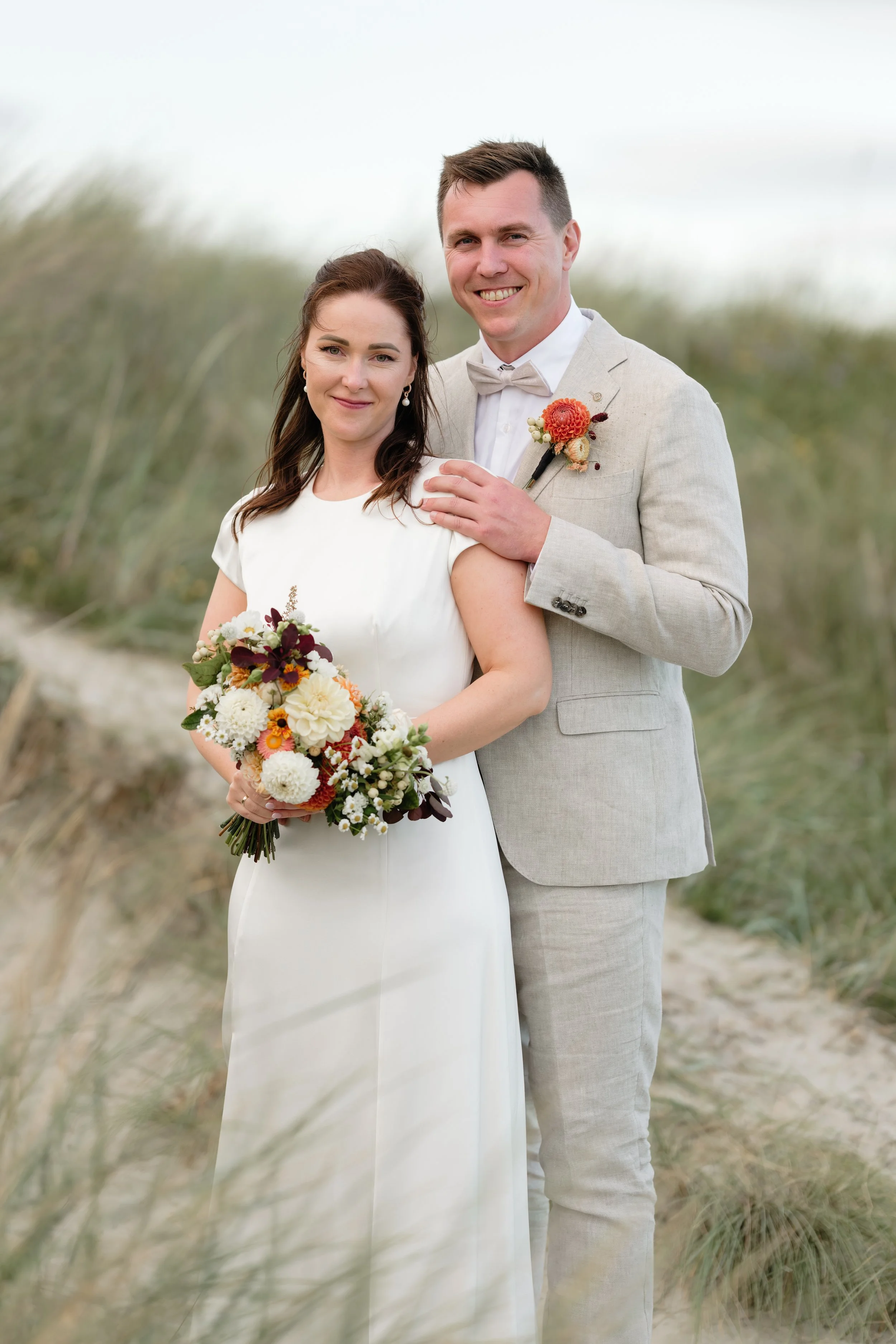 Et bryllupsbilde av et par på stranden med gress i bakgrunnen. Kvinnen holder en blomsterbukett, og mannen har en blomsterboutonnière.