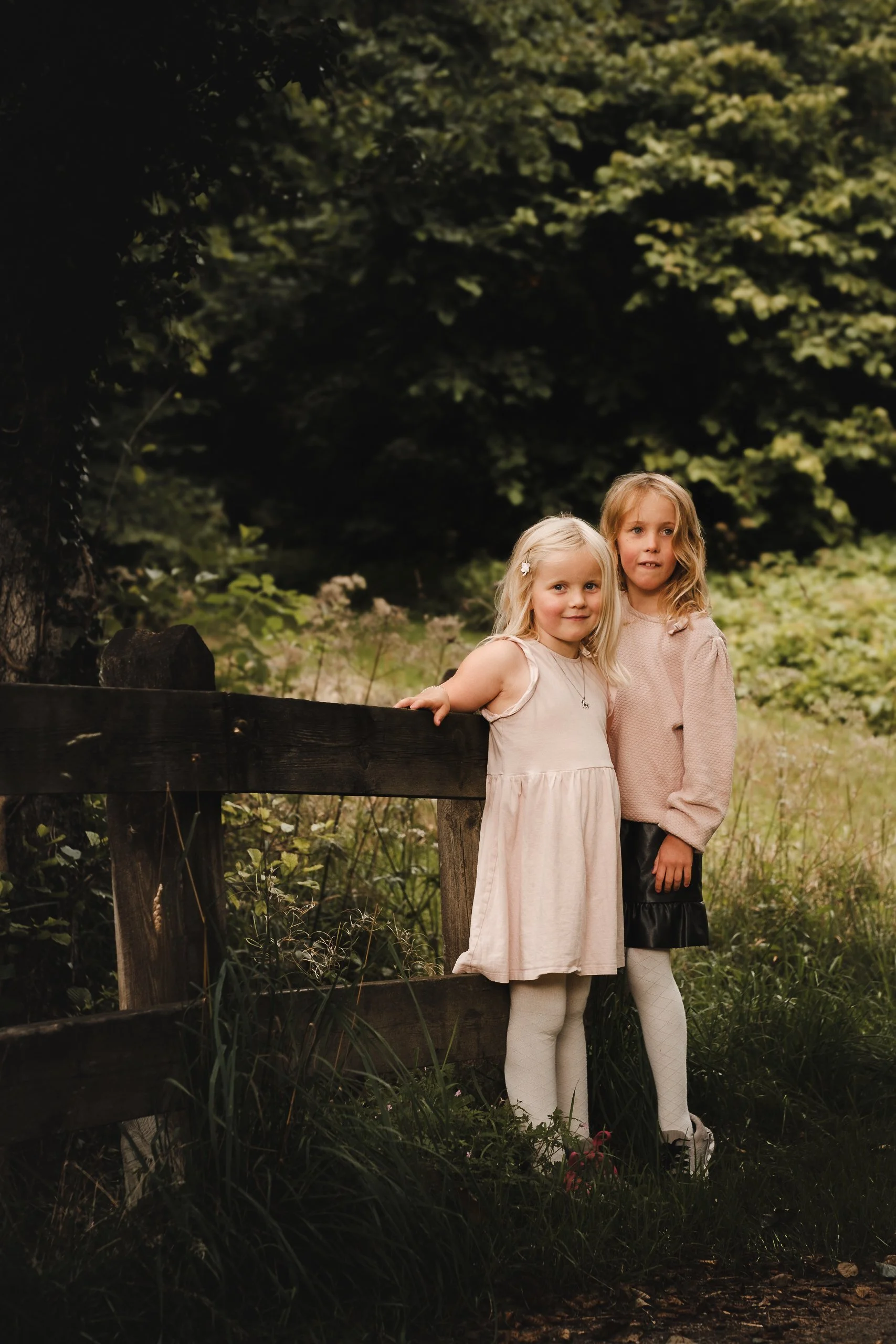 To blonde jenter i lyse kjoler står ved et tregjerde i en frodig skog.
