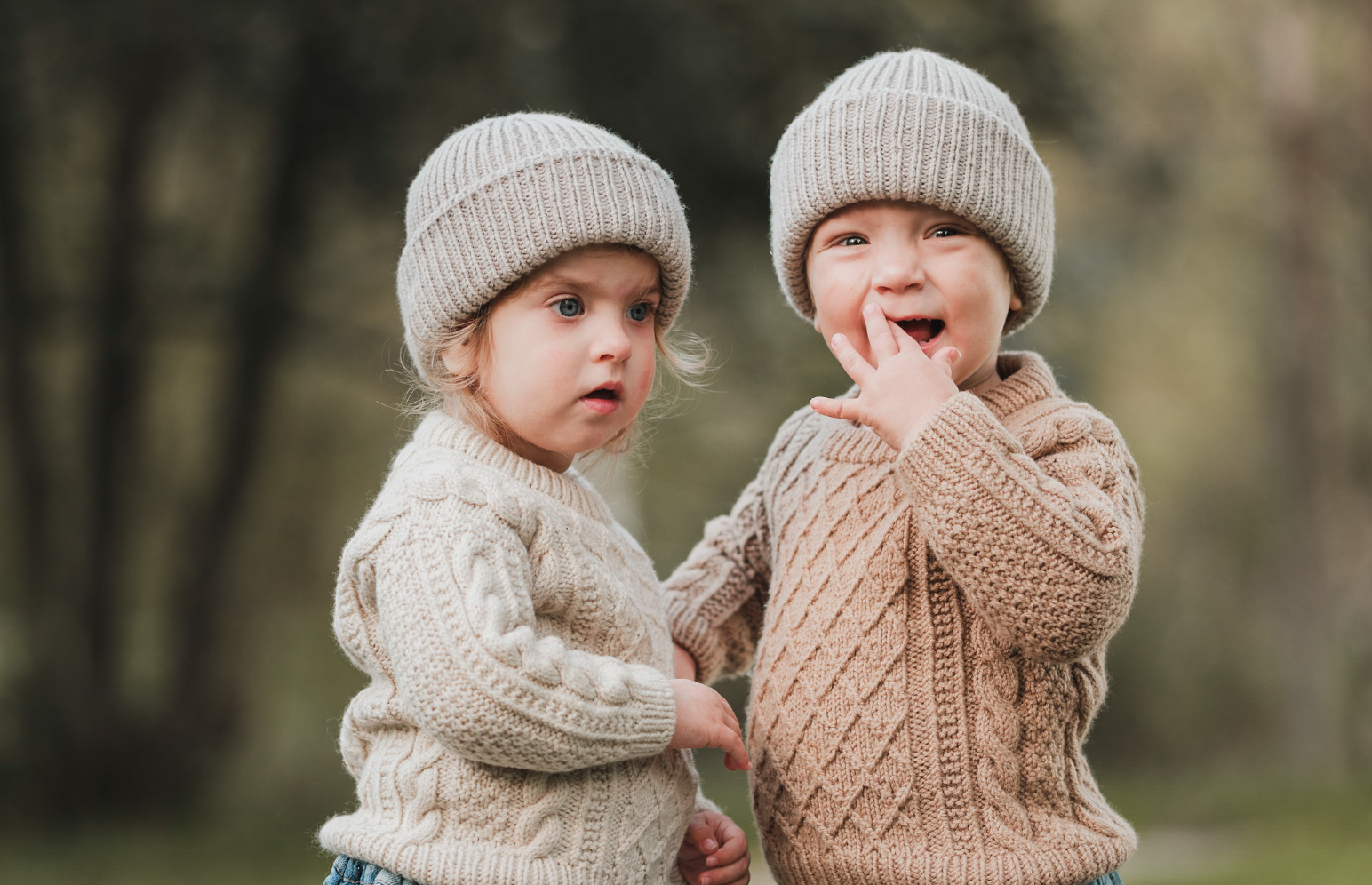 To unge barn, en gutt og ei jente, står utenfor i naturen iført beige strikkegenser og luer.