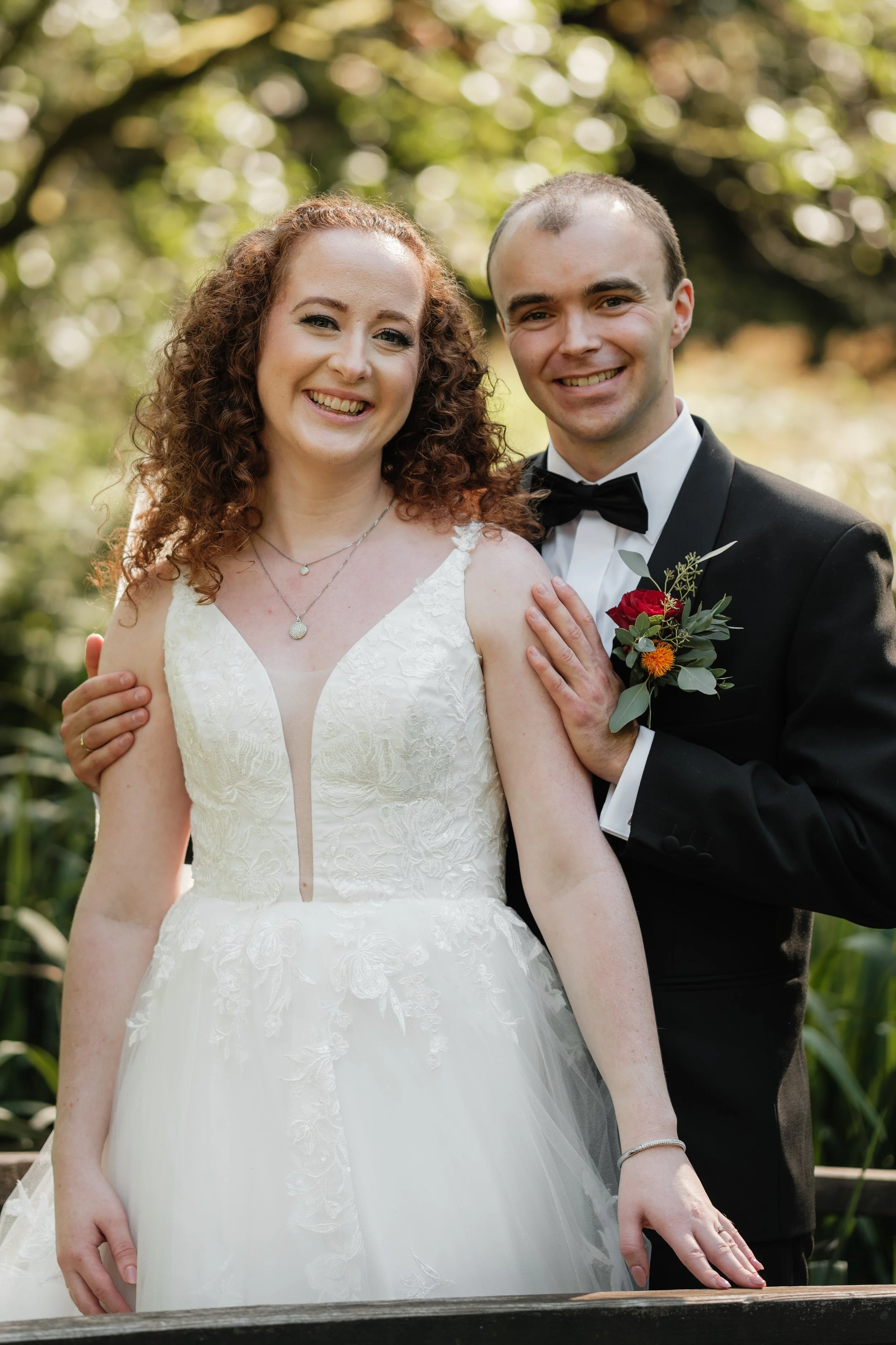 Et brudepar smiler under et tre i utendørs omgivelser. Bruden har rødt, krøllet hår og bærer en hvit brudekjole. Brudgommen har kort hår og bærer smoking med sløyfe og en blomsterbukett i lommen.