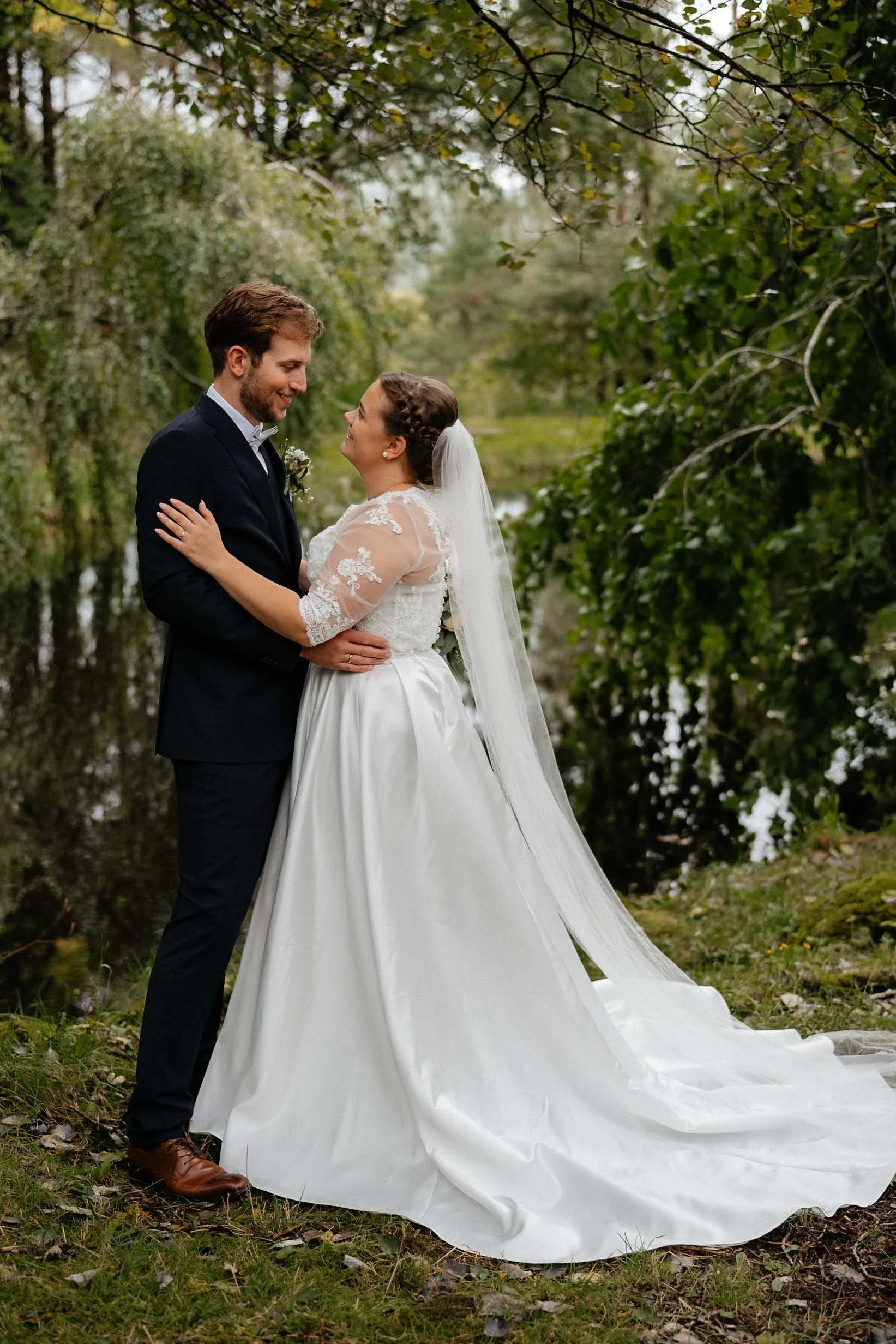 Nygift par omfavner hverandre i en frodig skog, med brud i hvit blondekjole og slør og brudgom i mørk dress.