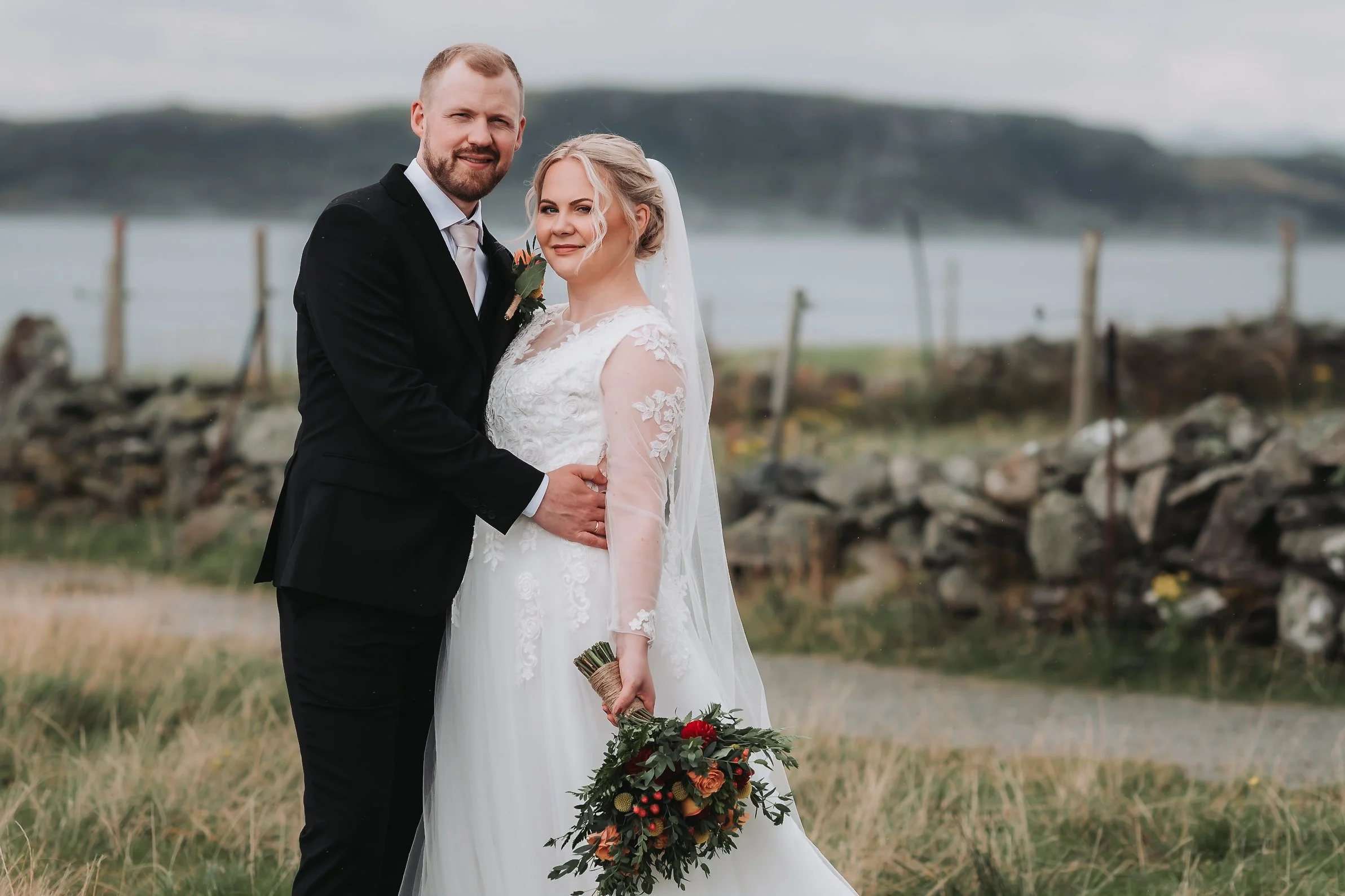 Et brudepar står ved vannet, iført brudekjole og dress, med landskap i bakgrunnen.