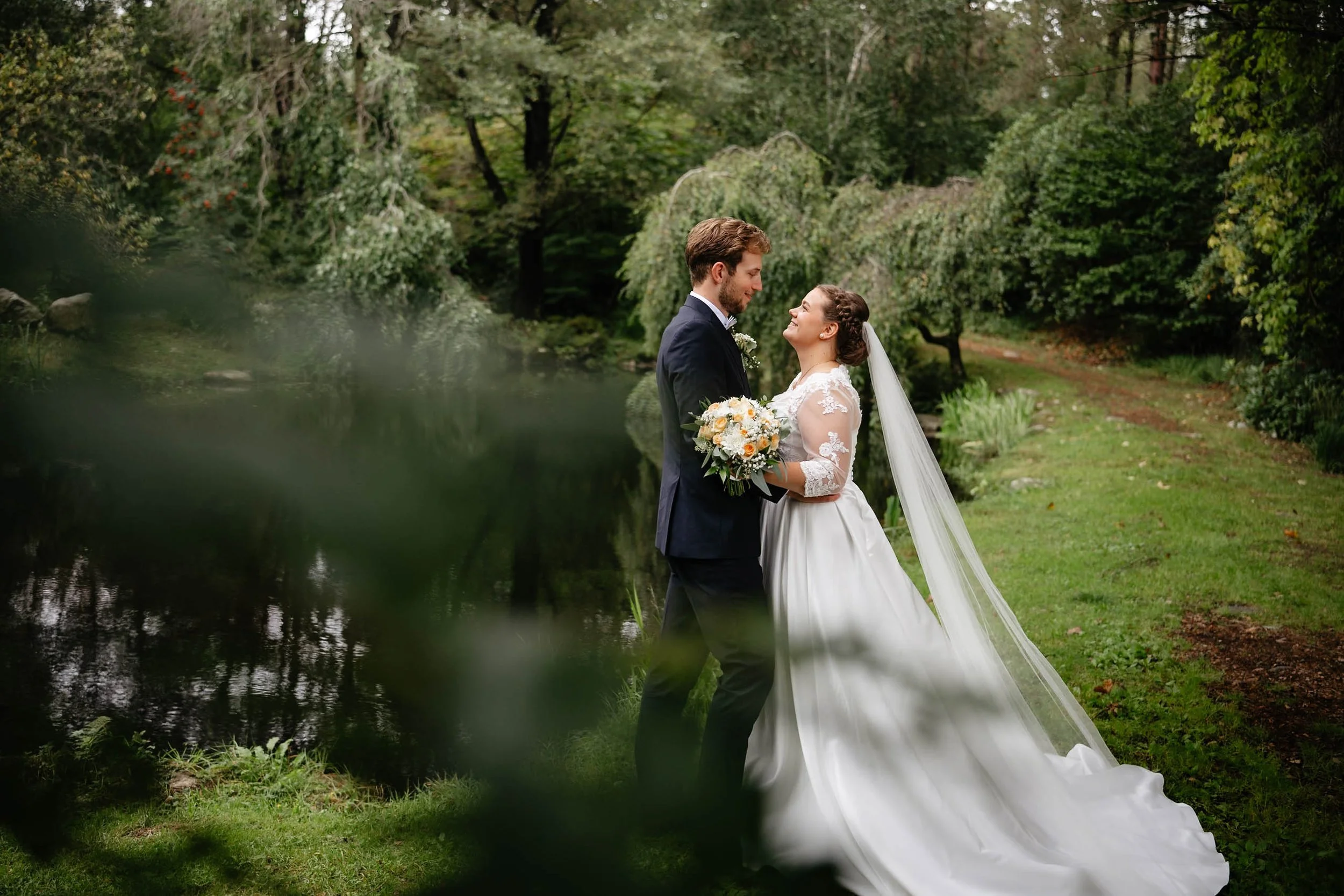 Brudepar i bryllupsklær ved en dam i en frodig park, med bruden holding en blomsterbukett.