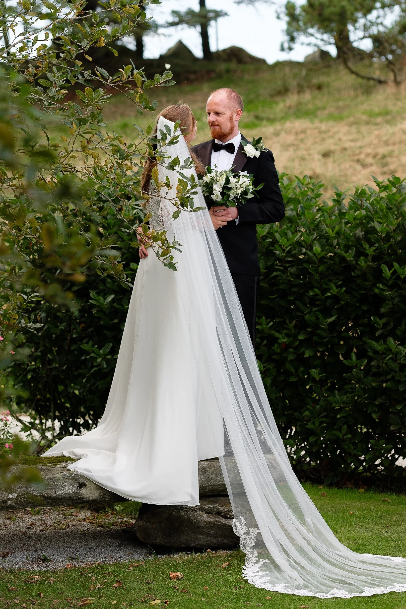 En brud og en brudgom står ansikt til ansikt i en hage, omgitt av grønne busker og trær, med brudens lange hvite kjole og slør.