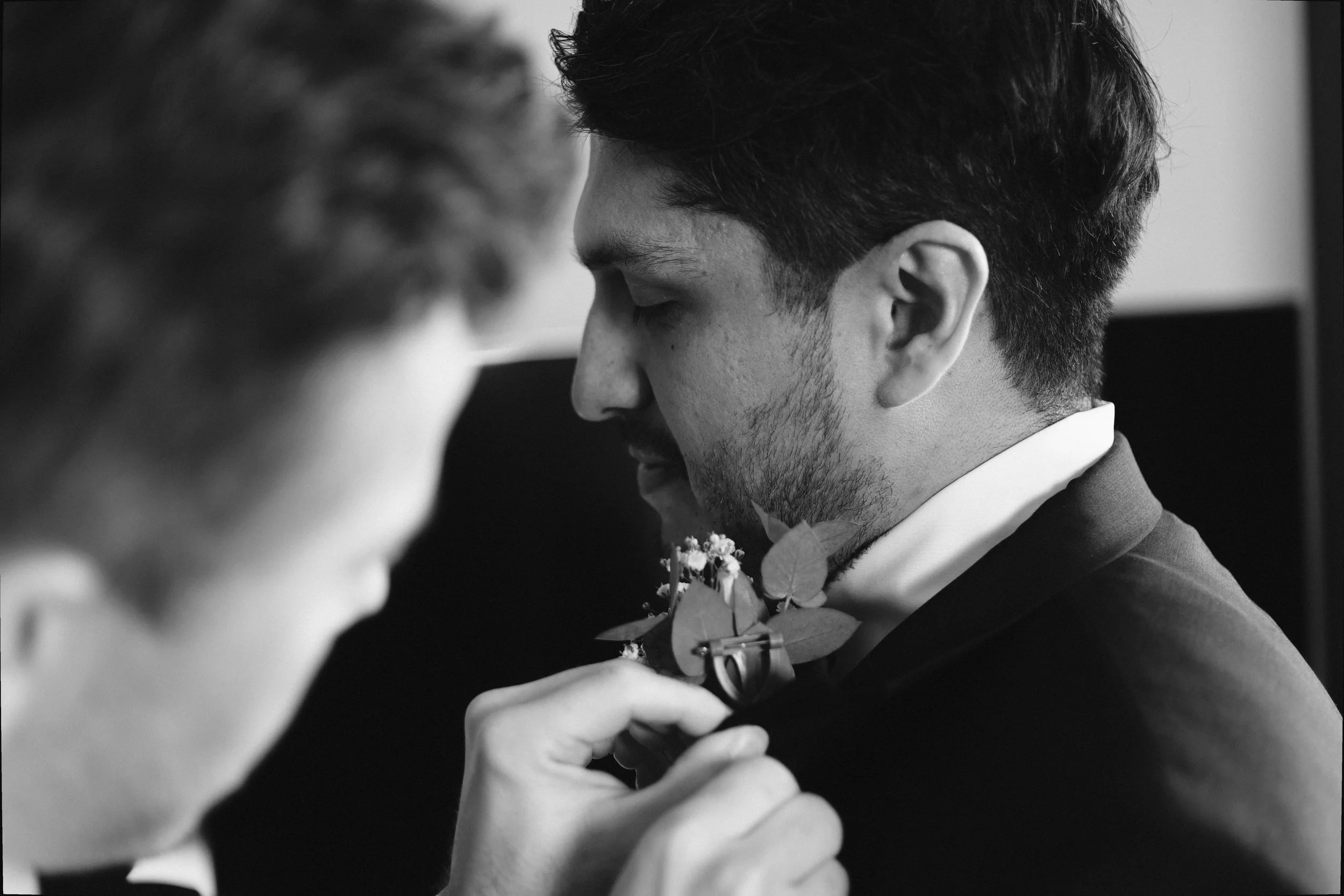 Svart-hvitt nærbilde av brudgom i dress med blomster, mens en person justerer boutonnièren hans.