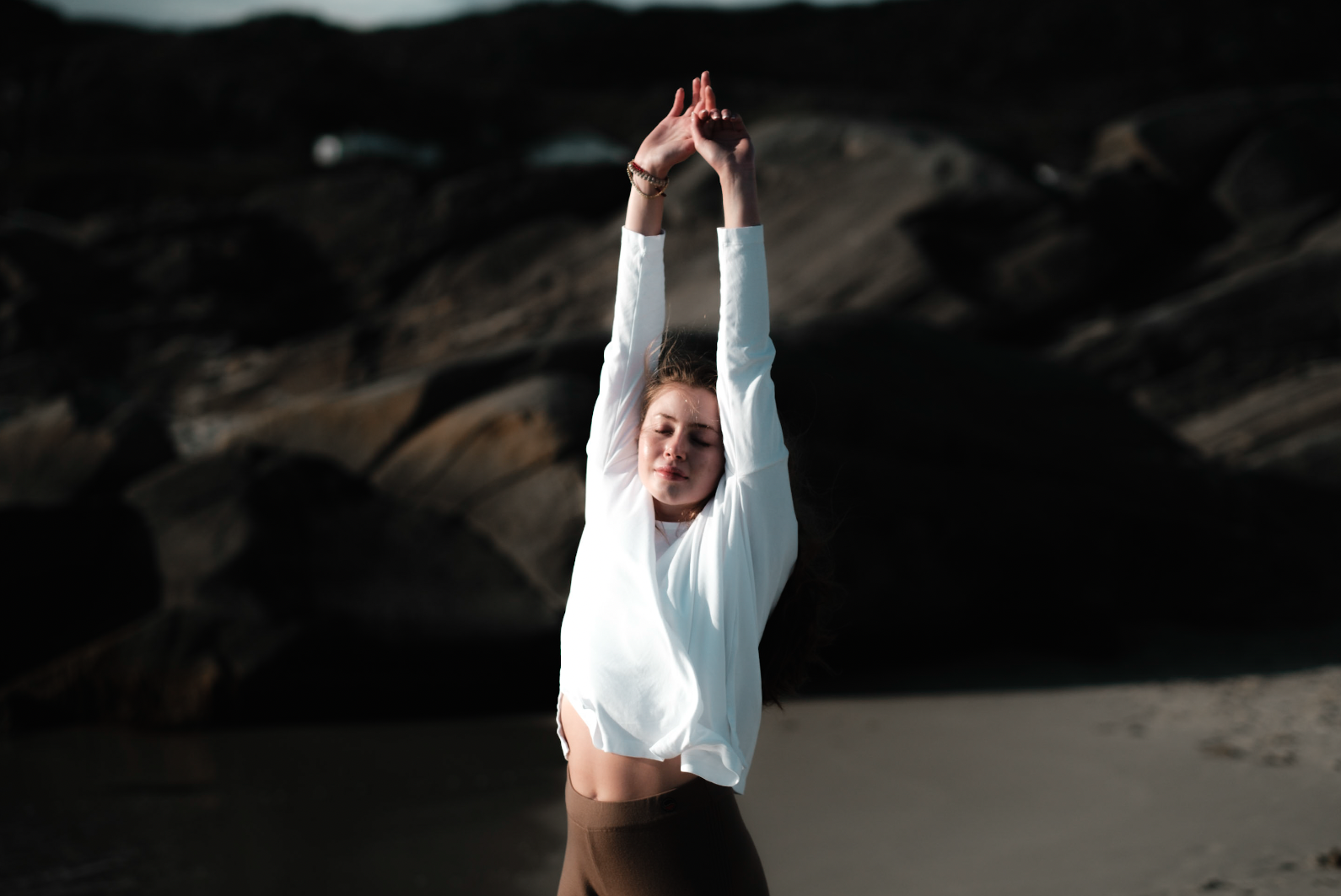 Kvinne med lang hår i hvit topp og brune bukser står på stranden med armene hevet over hodet, solnedgang eller skumring i bakgrunnen.
