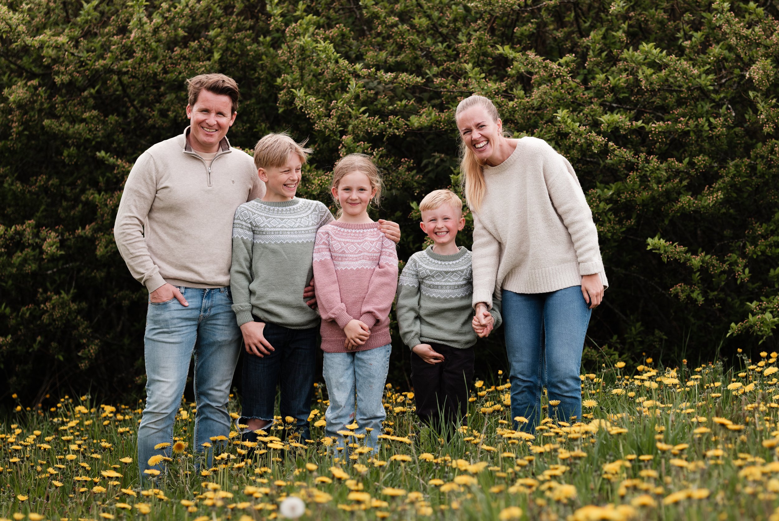 En familie av seks personer, to voksne og fire barn, står i en blomstereng med gule blomster og grønne busker i bakgrunnen. De smiler og holder hender.