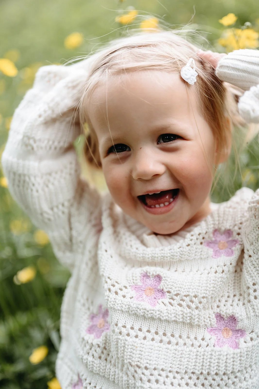 Smilende småbarn i hvit strikkegenser med lilla blomster, omgitt av gule blomster i en hage.