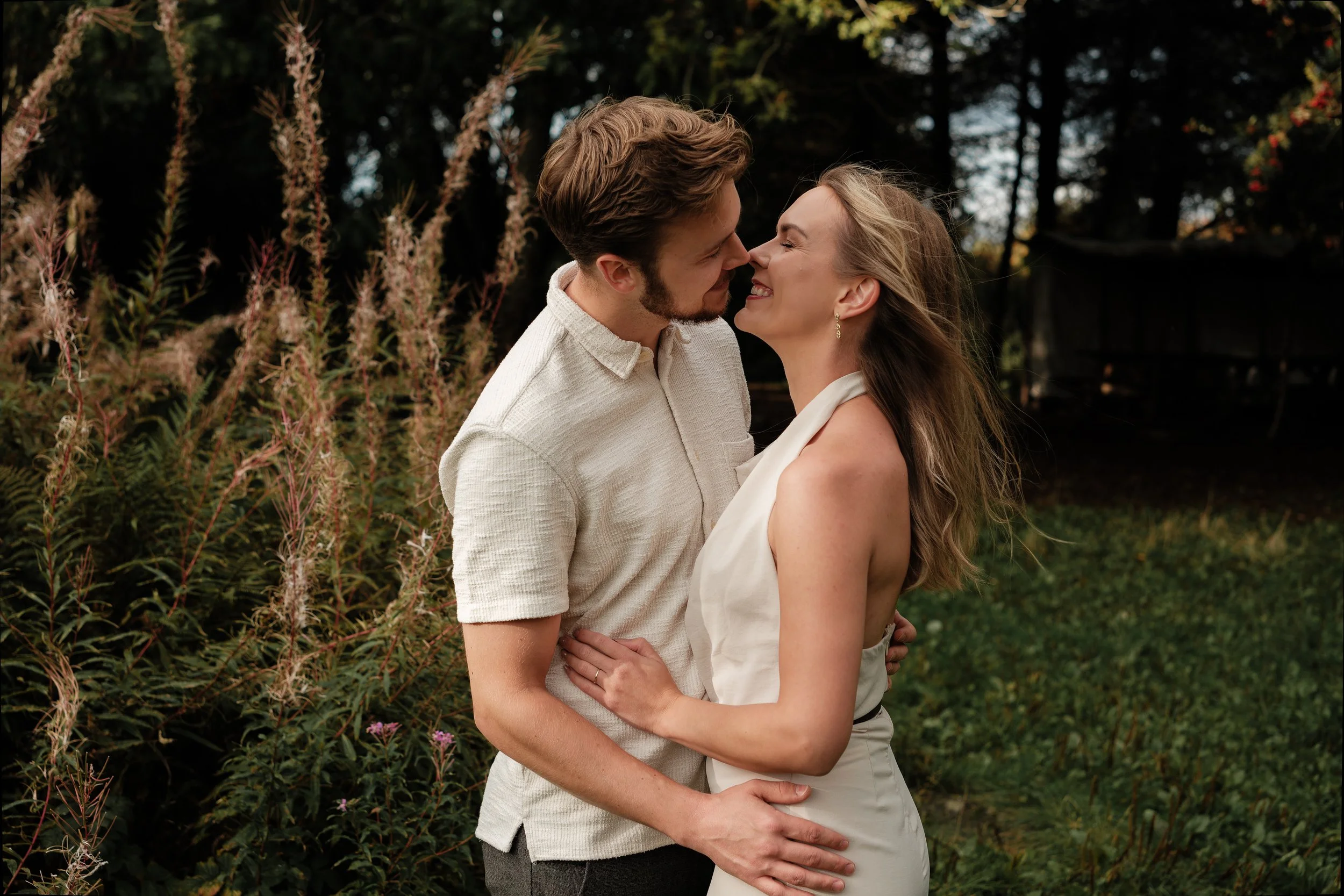 Et kjærlighetspar står nært sammen i en hage, smiler og nikker mot hverandre, omgitt av grønne planter og blomster.